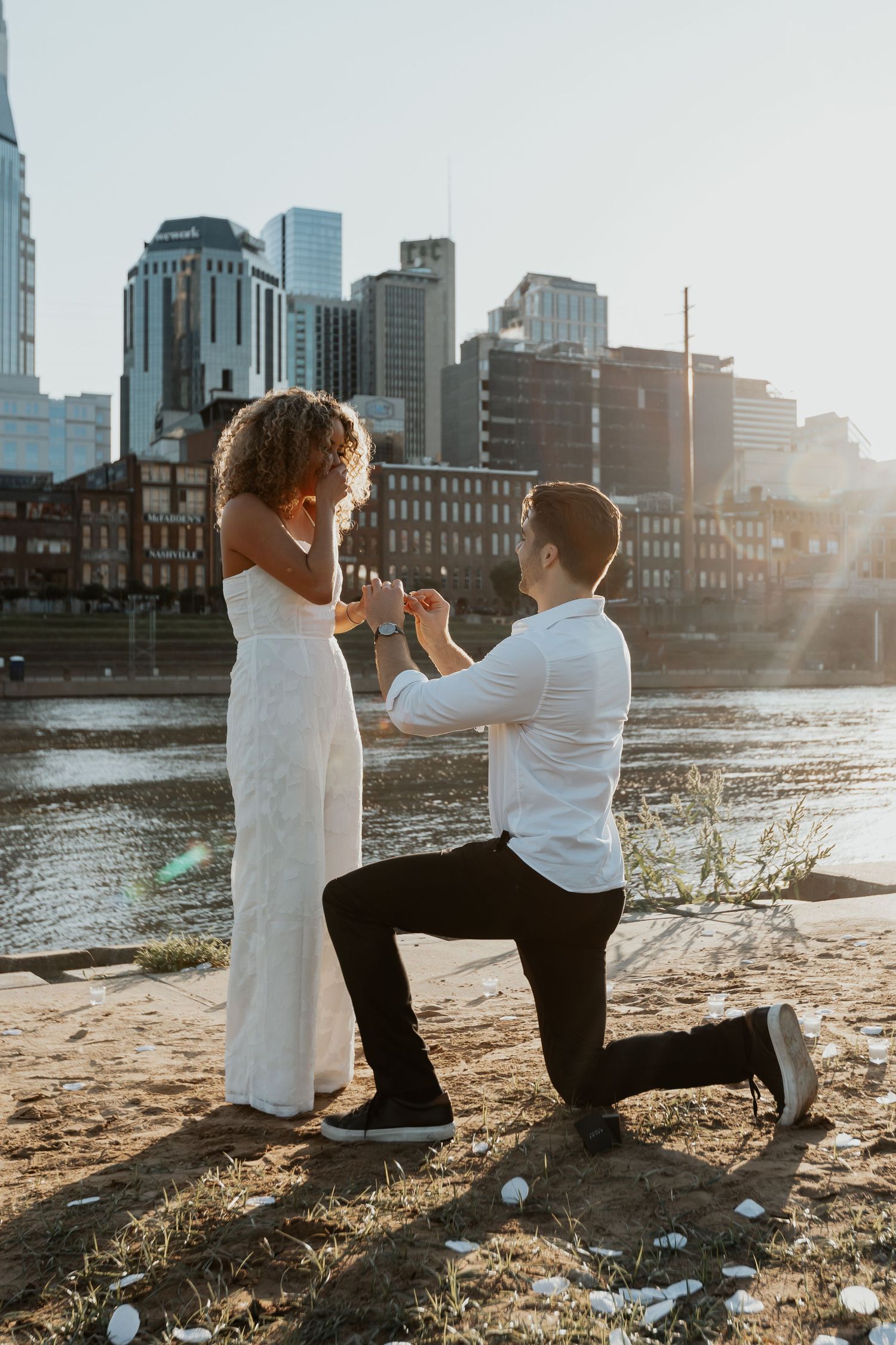 Man proposes on one knee to his girlfriend on a riverbank with a city skyline at sunset.