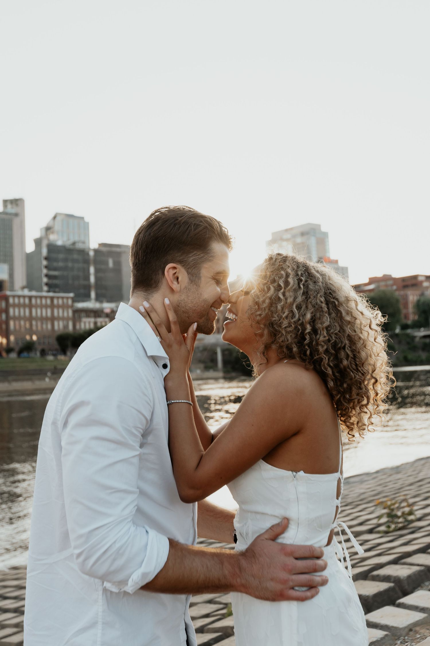 Couple shares a romantic almost-kiss embrace near a waterfront with a city skyline at golden hour.