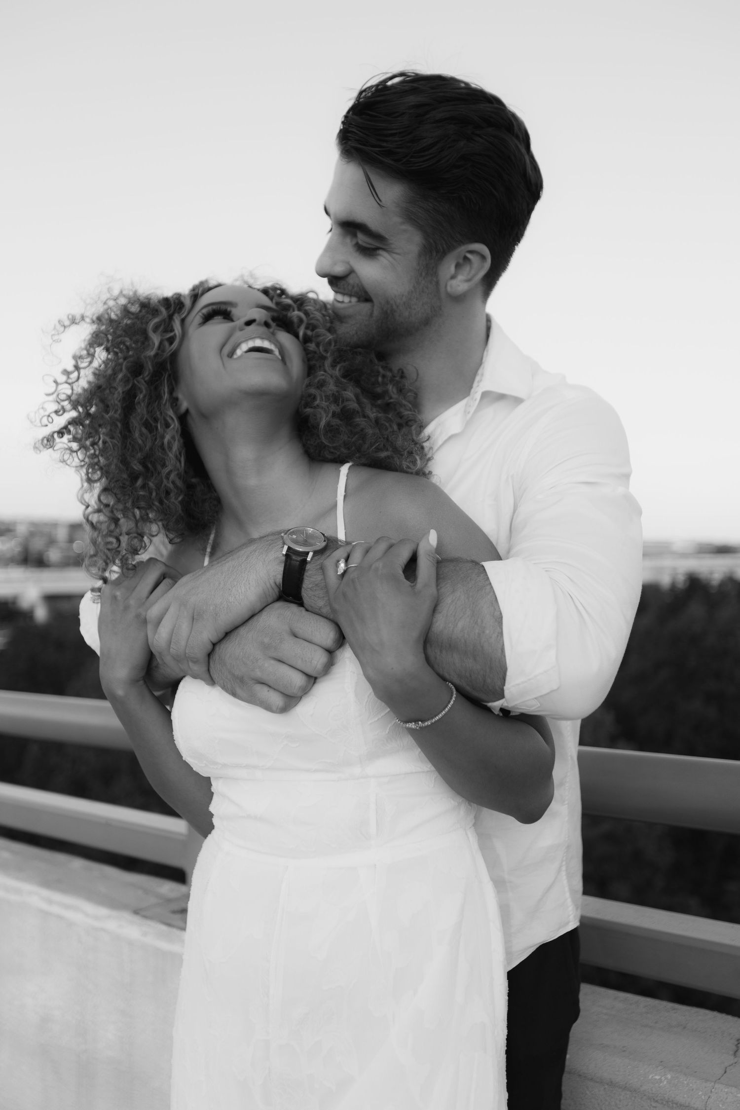 Black and white photo of a smiling couple embracing on a rooftop overlooking a city skyline.