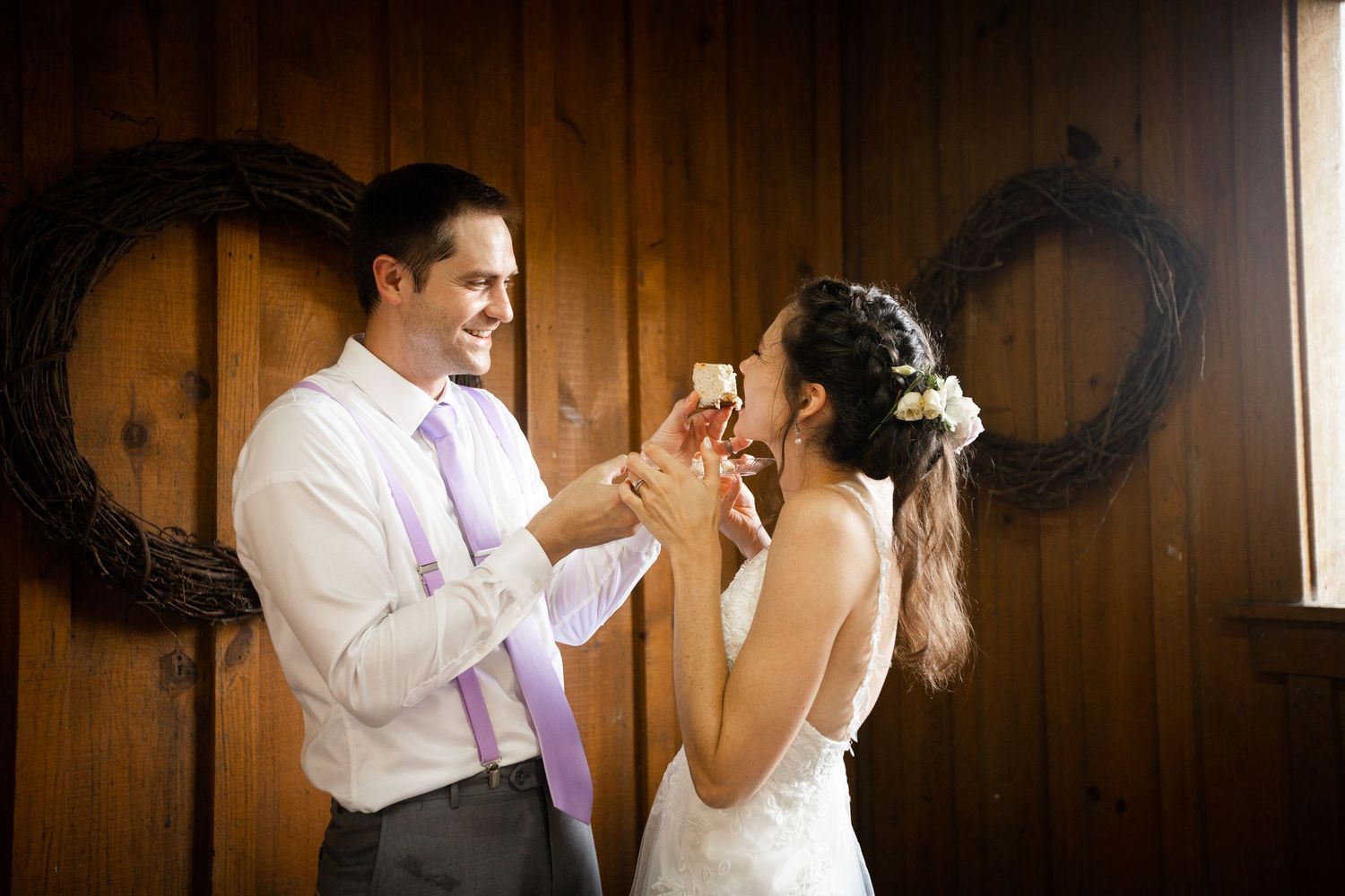 Bride and groom sharing wedding cake in rustic barn decorated with wreath and lavender accents.
