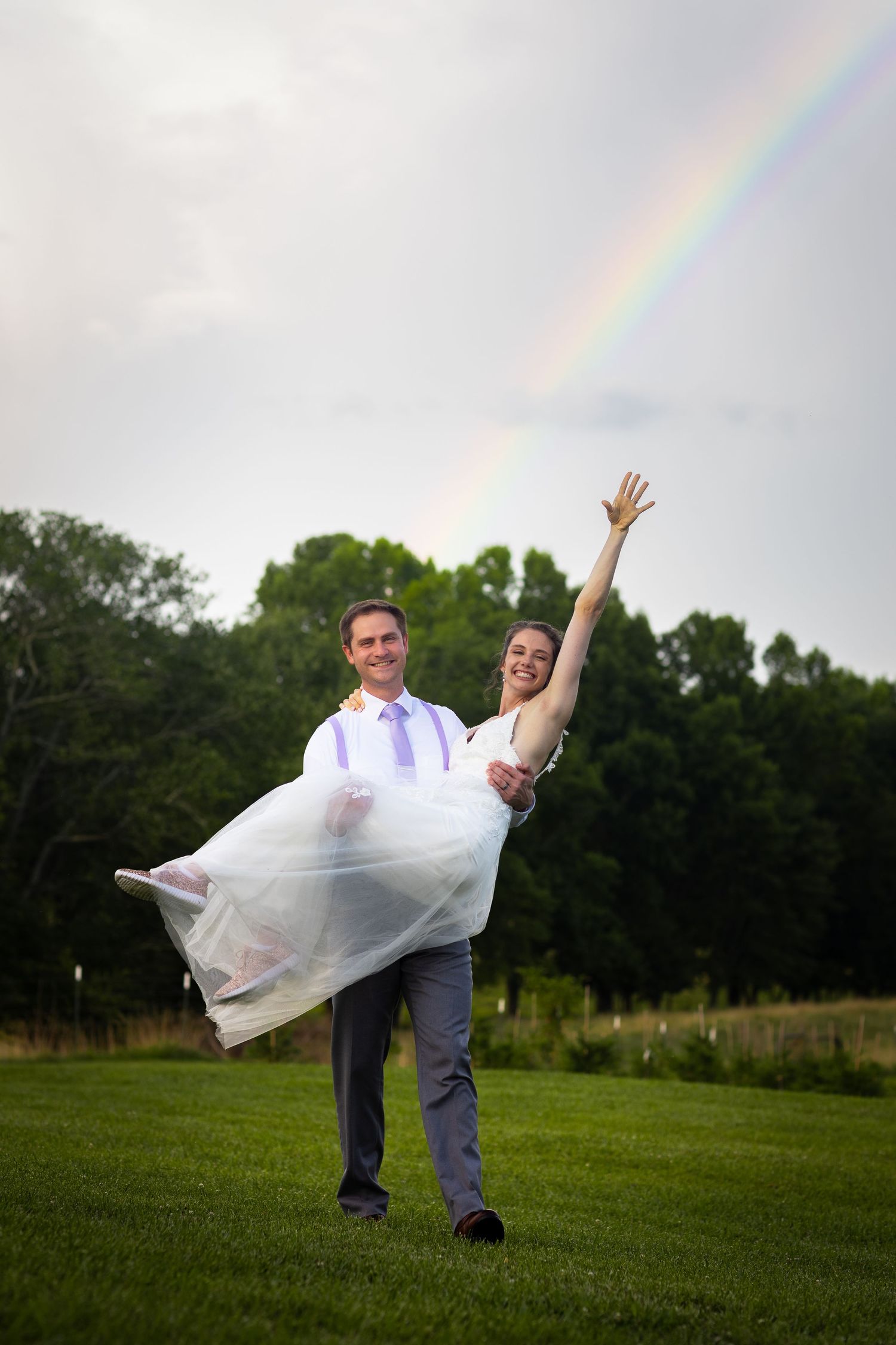 Groom carries joyful bride in white dress under a rainbow in green outdoor wedding setting.