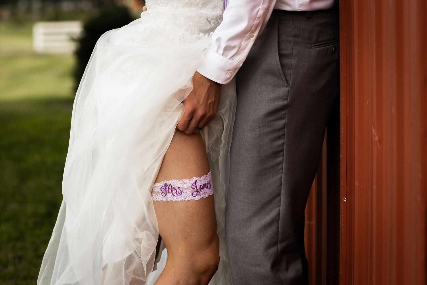 Bride lifting wedding dress to reveal a purple Mrs. garter on her leg while standing next to groom in gray suit.