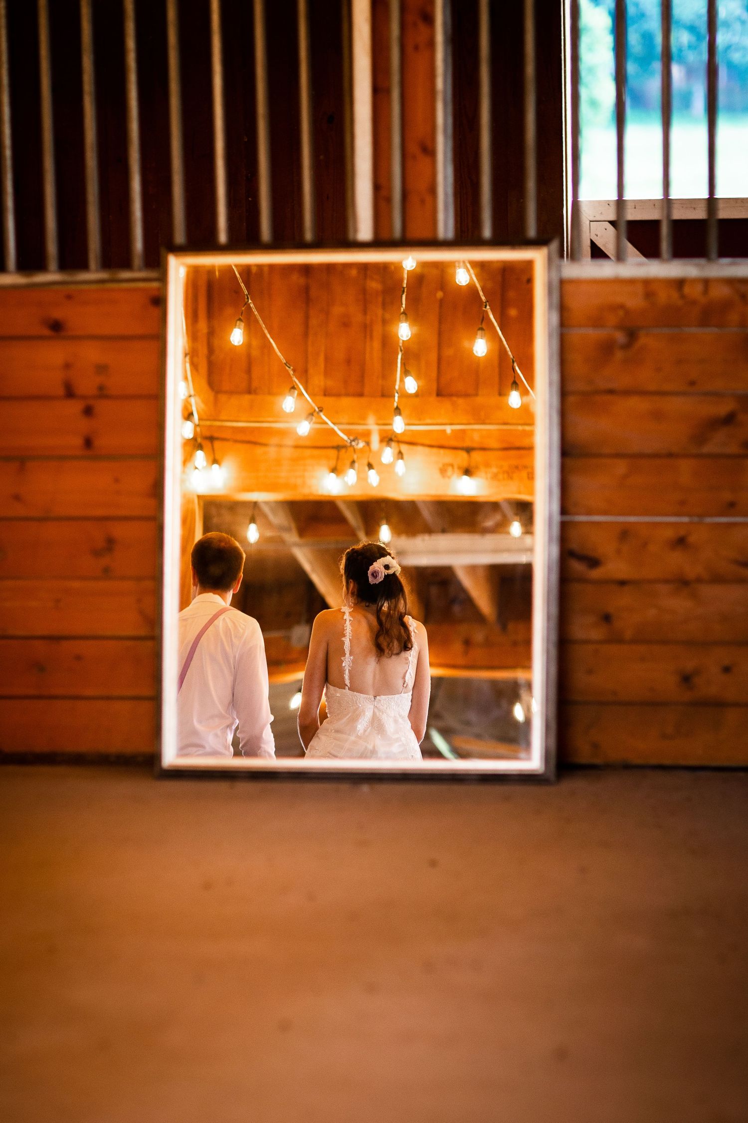Wedding couple reflected in ornate mirror inside rustic barn decorated with warm string lights.