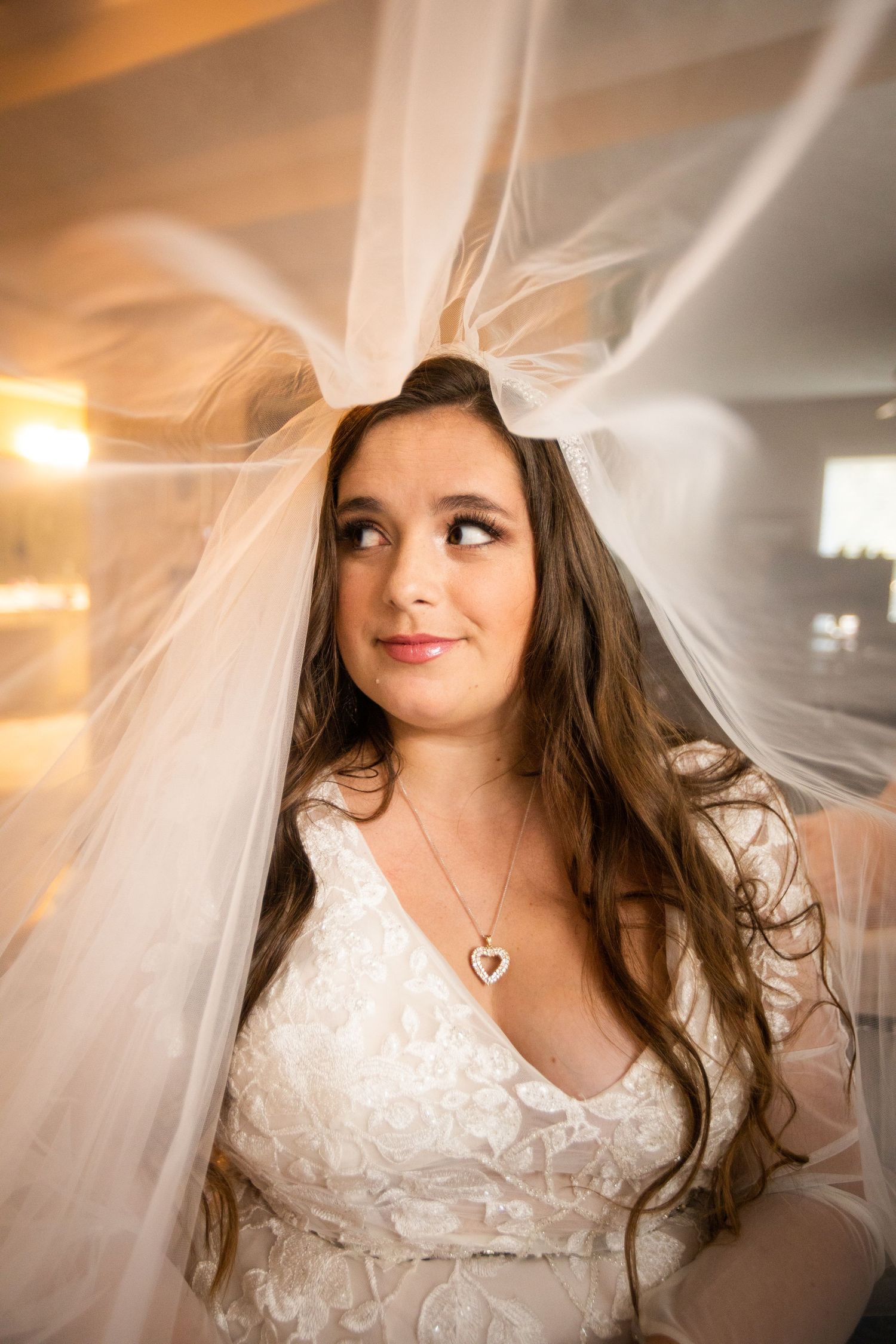 Beautiful bride with long brown hair wearing lace gown and heart necklace under flowing white veil.