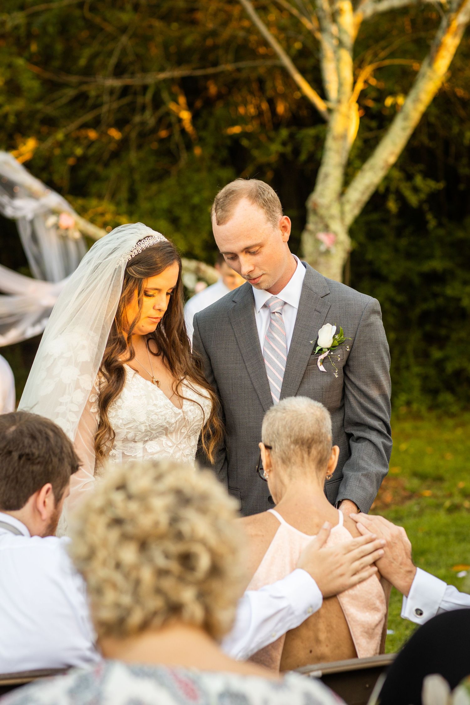 Bride and groom bowing heads in prayer with guests at outdoor autumn wedding ceremony.