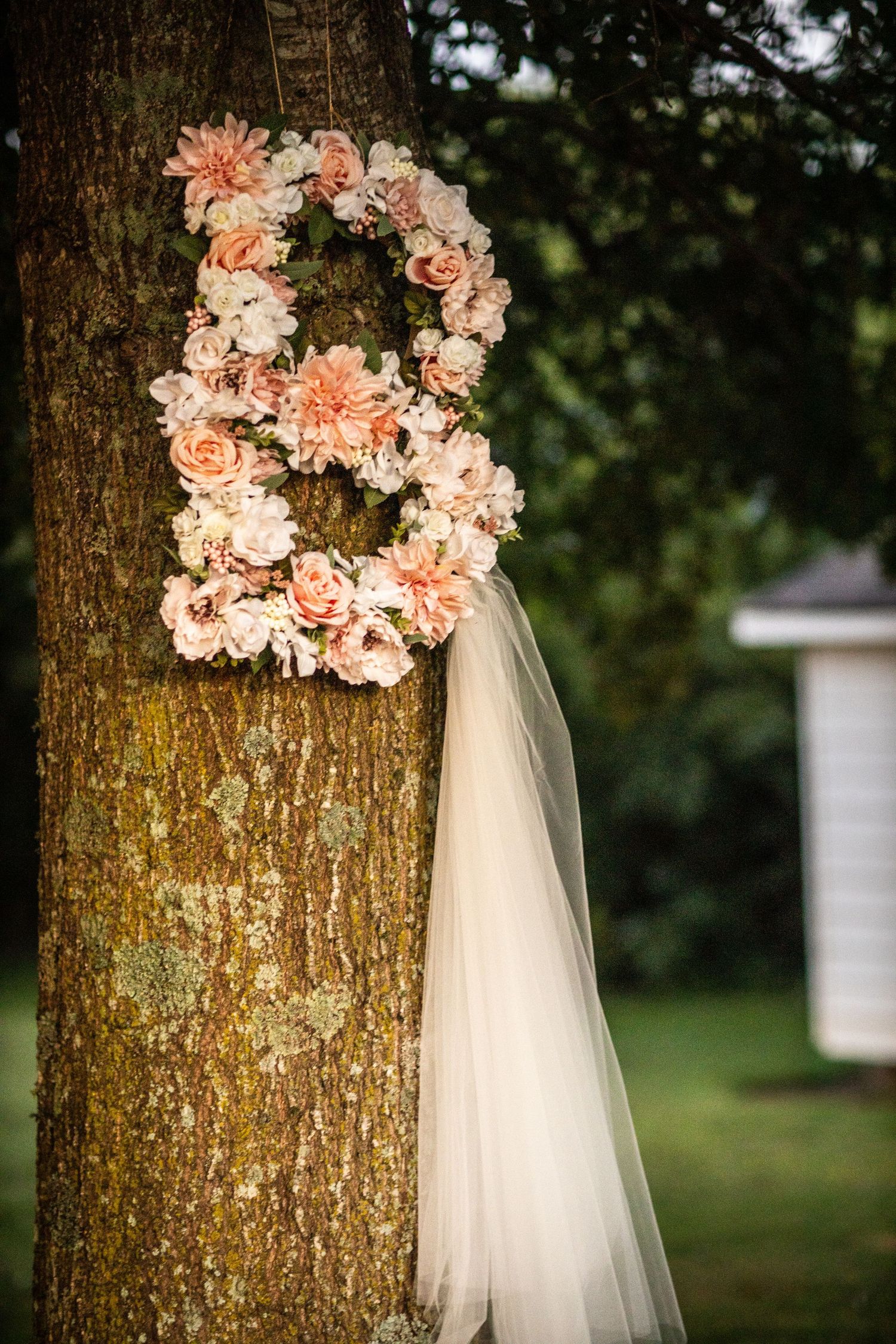 Floral letter B decoration made of peach roses and dahlias with white veil attached to tree.