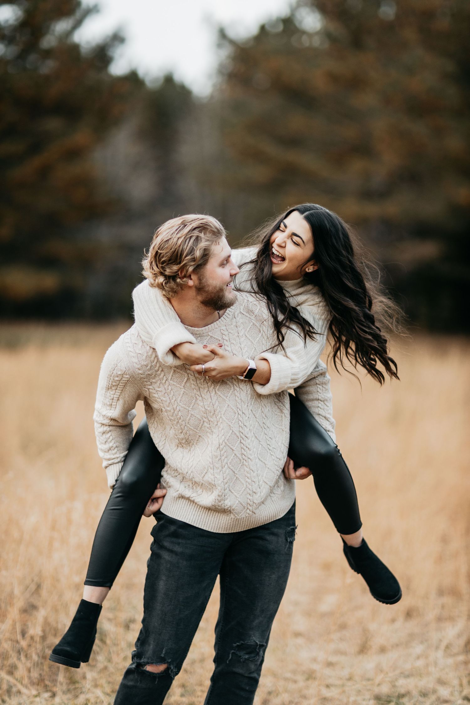 Young couple laughing in autumn field, man giving woman piggyback ride wearing cozy cream sweaters.