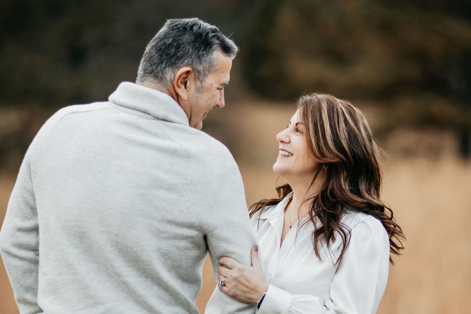 Happy middle-aged couple smiling at each other in golden autumn field wearing casual sweaters.