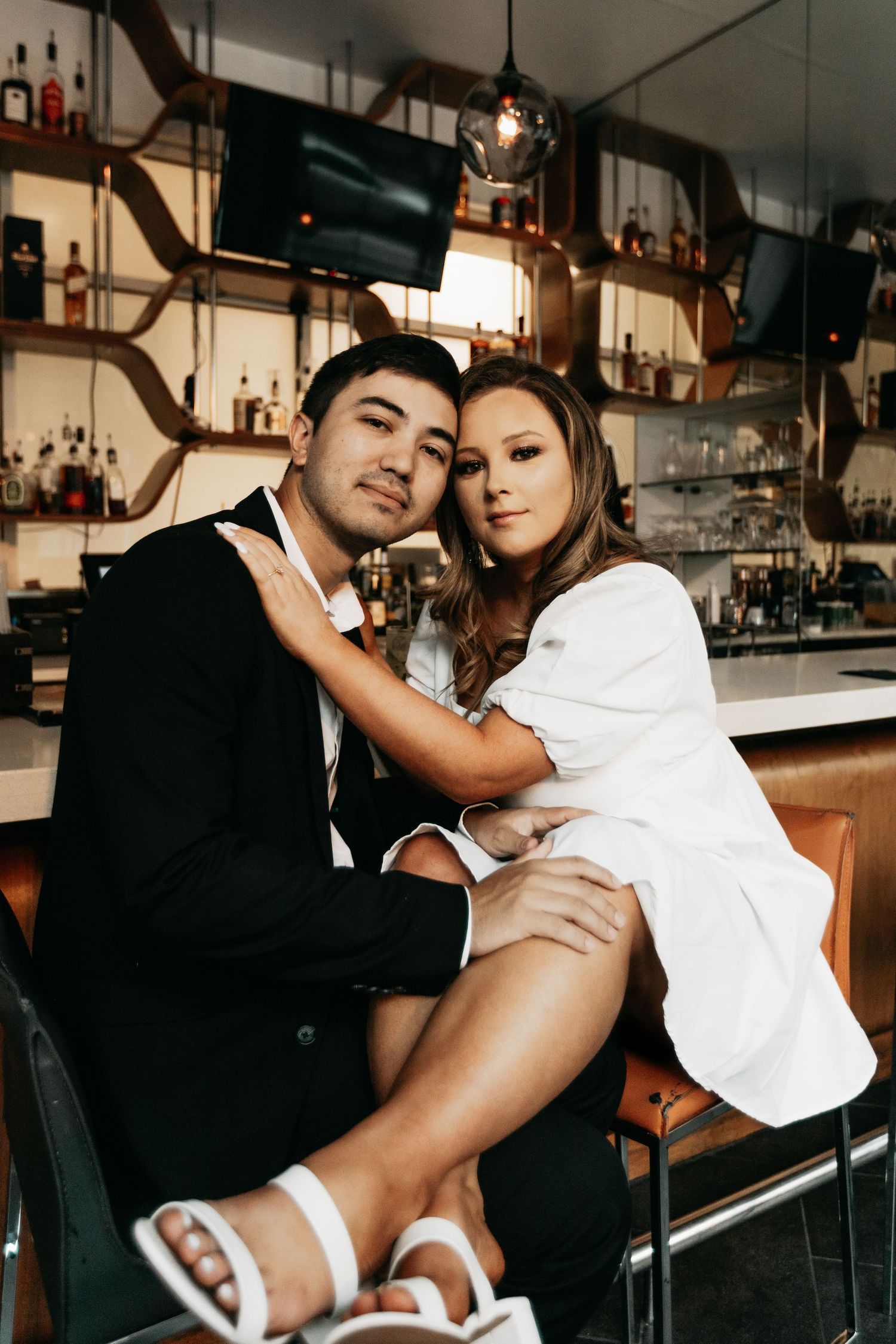 Stylish couple posing together at upscale bar, woman in white dress sitting on mans lap.
