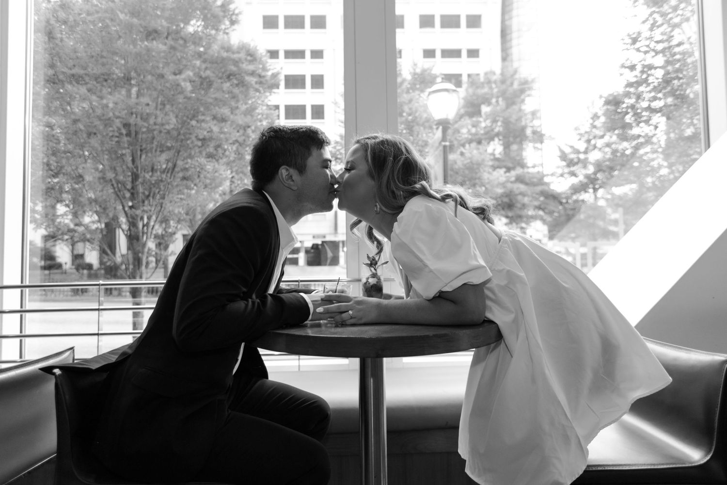 Black and white photo of a couple sharing a romantic kiss at a cafe table with city buildings in the background.
