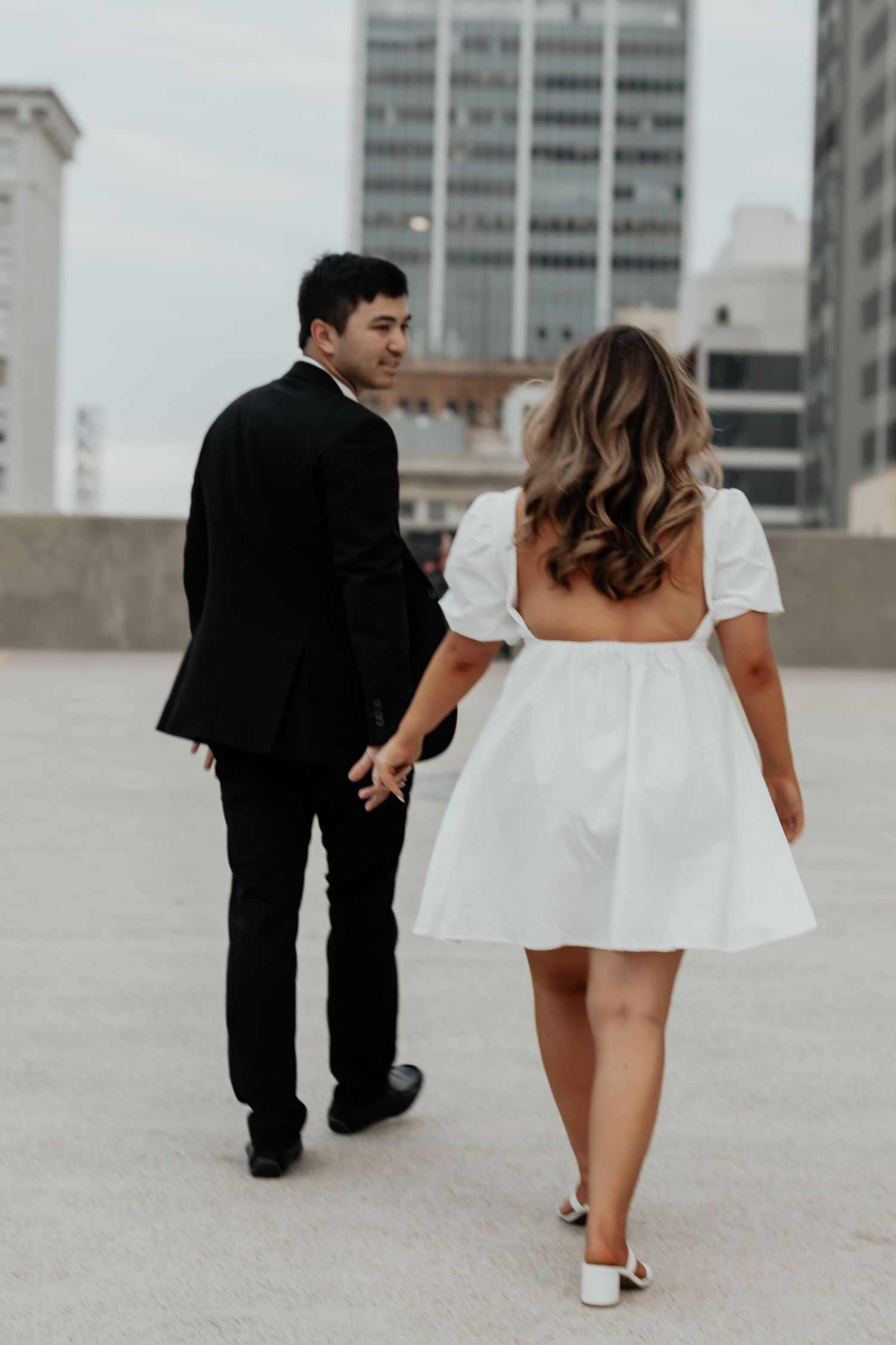 Couple holding hands walking on a rooftop, man in black suit and woman in white puff-sleeve mini dress.