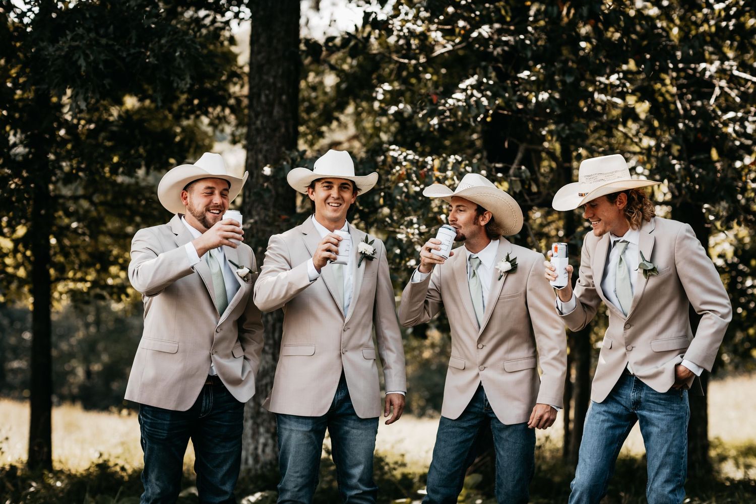 Four groomsmen in tan blazers, jeans, and cowboy hats toasting outdoors in a forest.