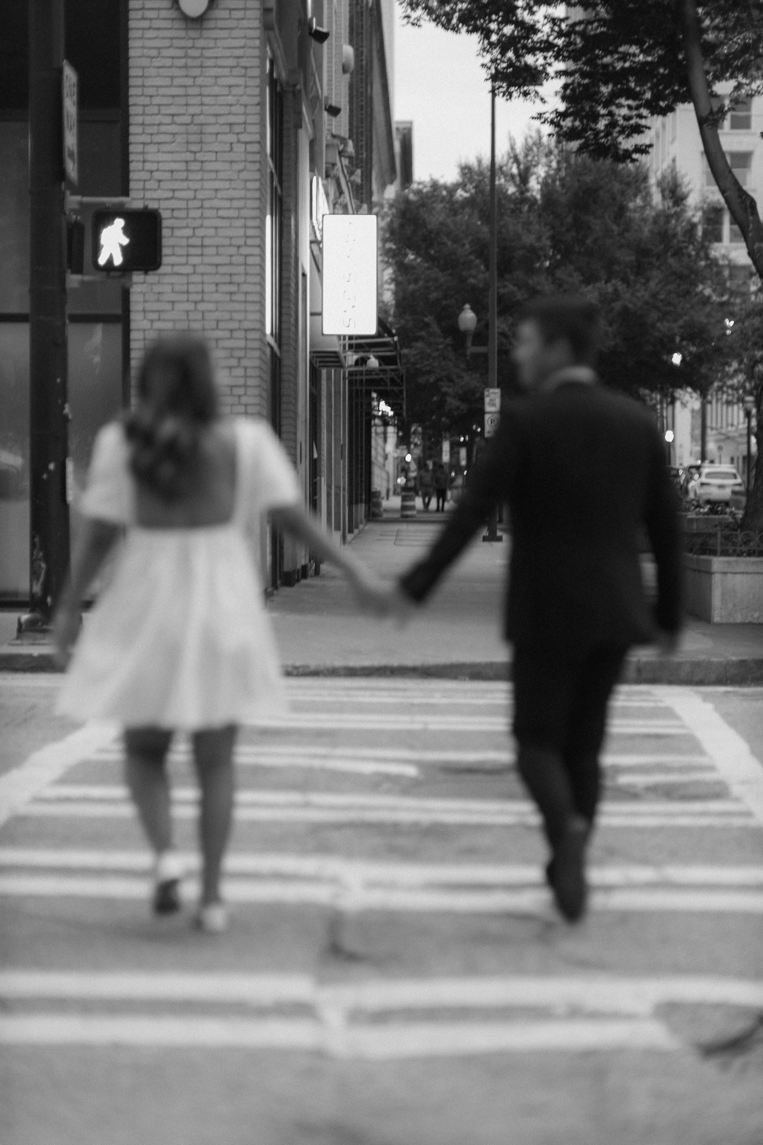 Black and white photo of a couple holding hands while crossing a city street crosswalk.