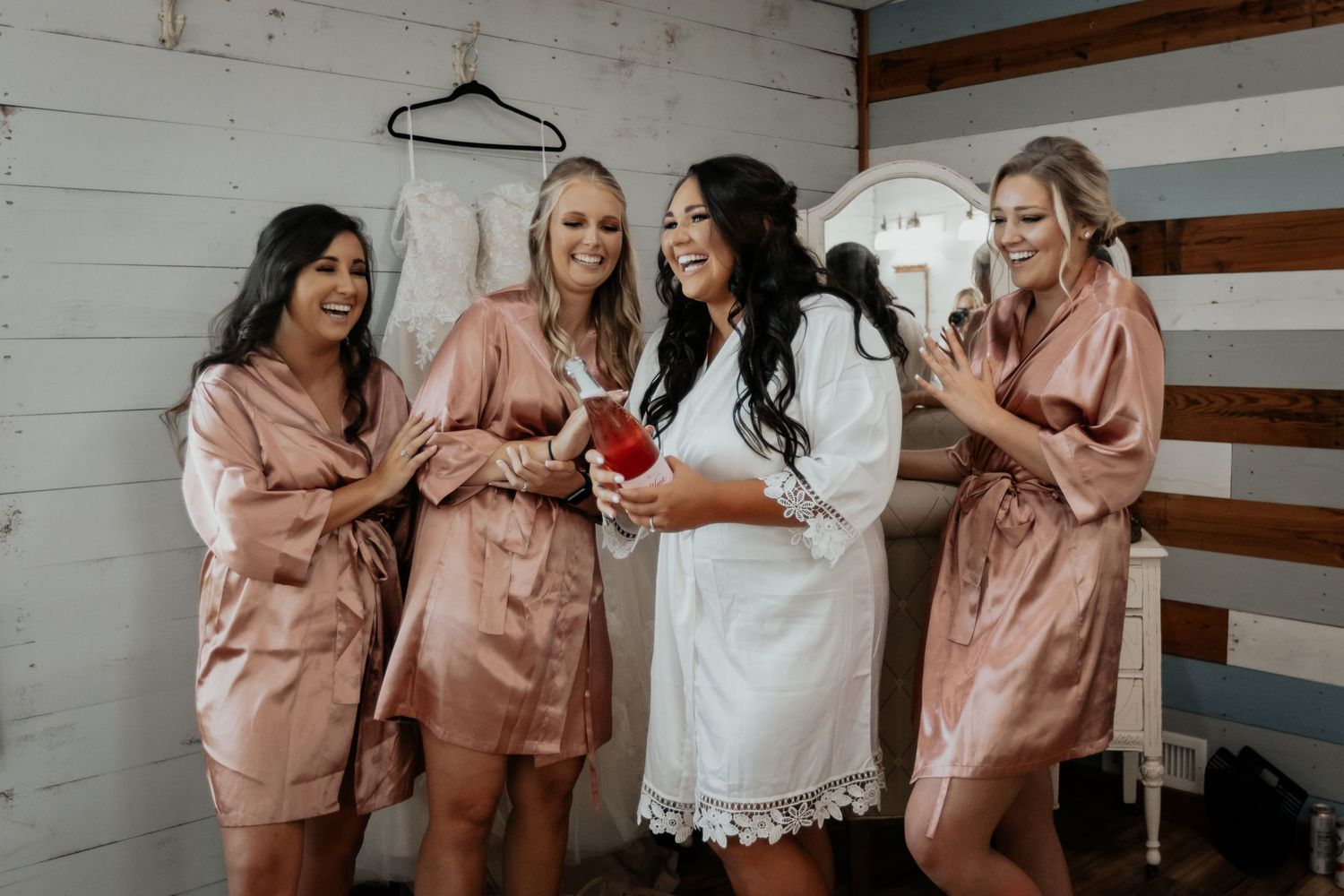 Bride and bridesmaids in satin robes laughing and celebrating together before the wedding.