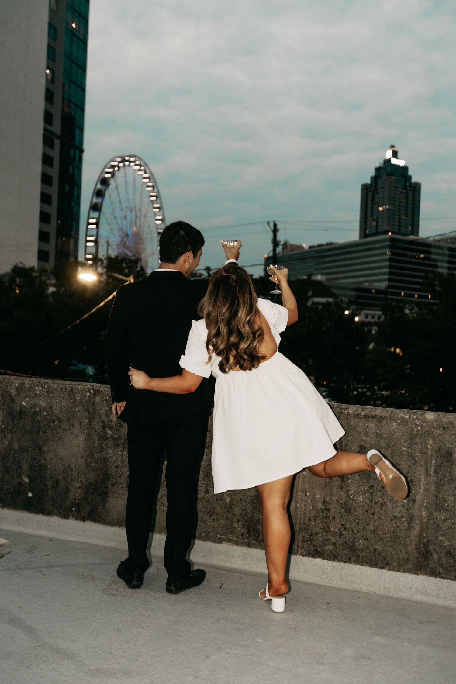 Couple embracing on a rooftop at dusk with a Ferris wheel and city skyline in the background.