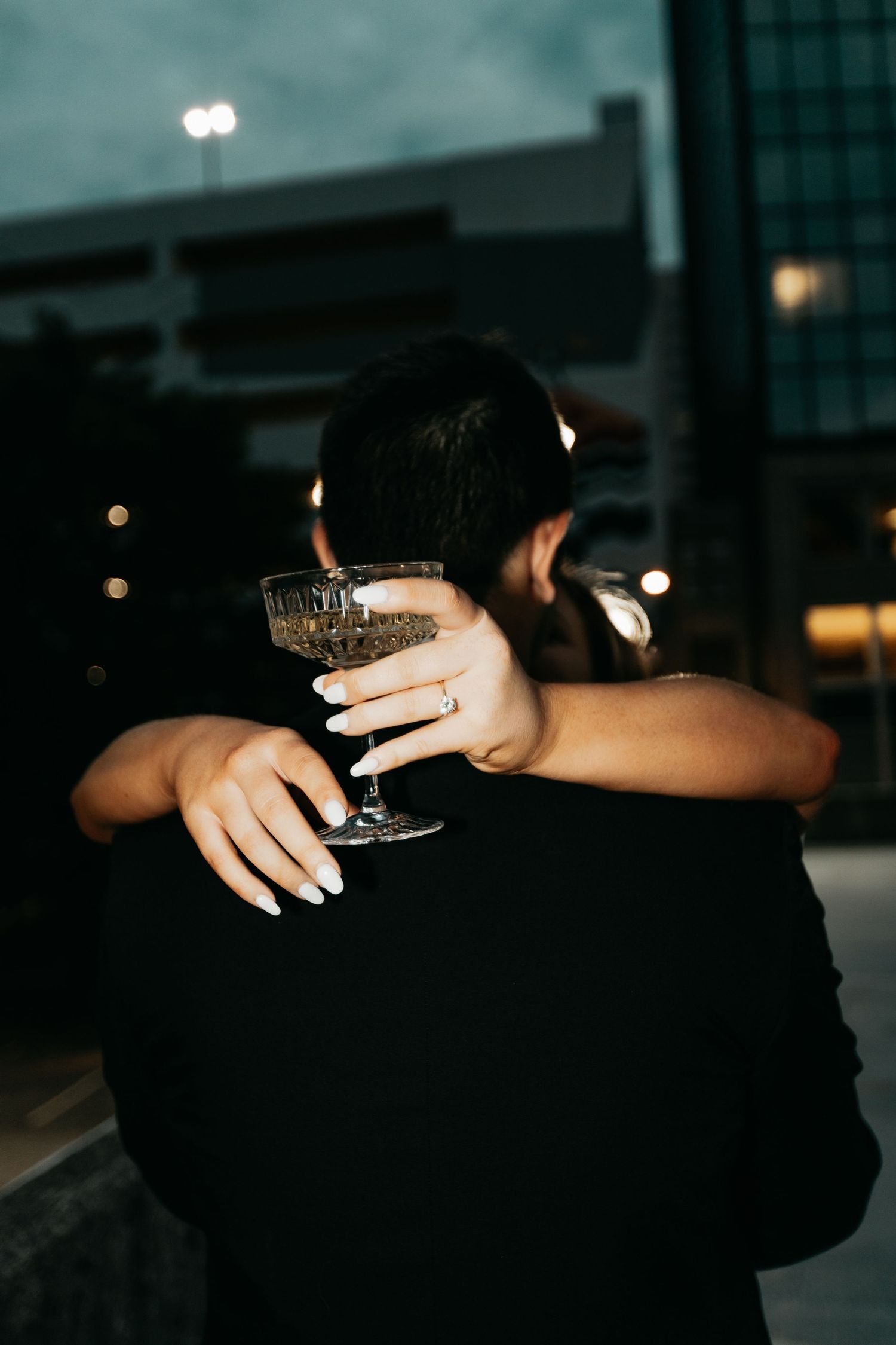 Close-up of a woman hugging a man from behind while holding a champagne coupe glass with an engagement ring visible.