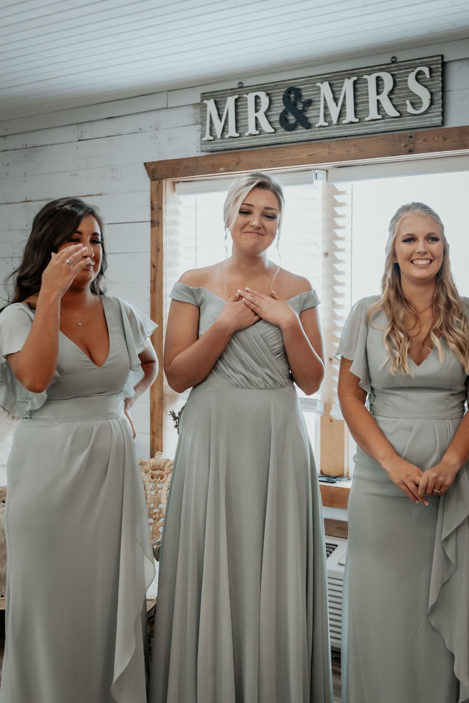 Three bridesmaids in sage green dresses smiling emotionally under a Mr & Mrs sign.