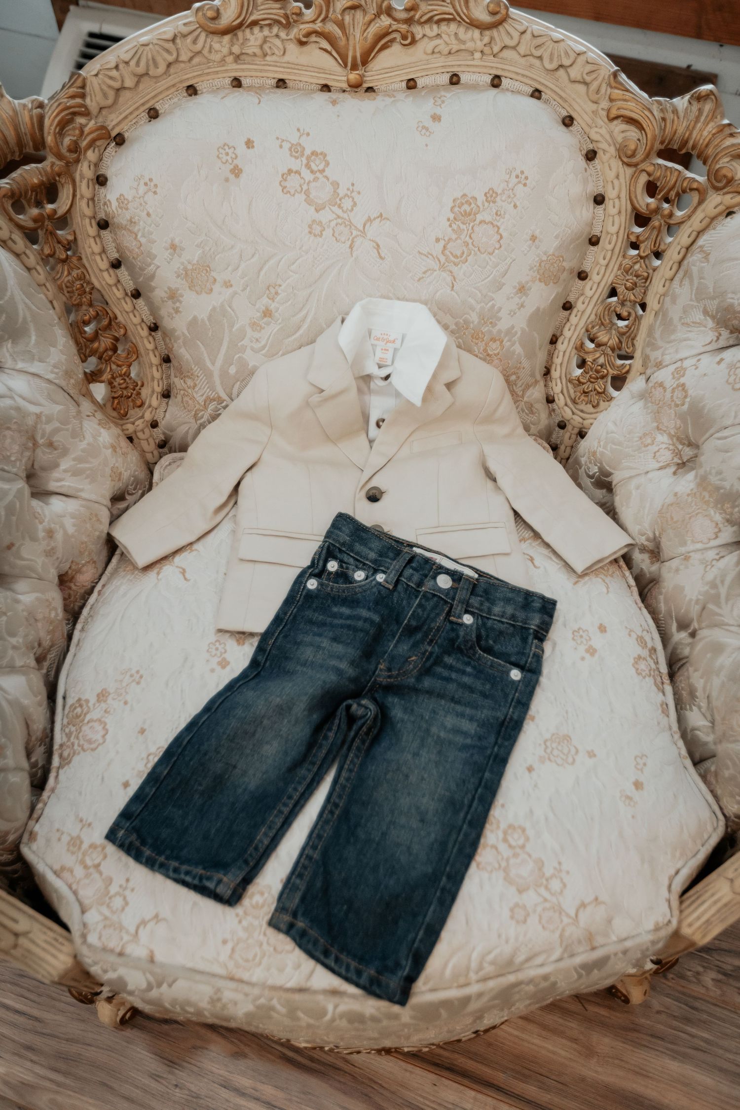 Small boys beige blazer and jeans outfit laid out on an ornate vintage chair.