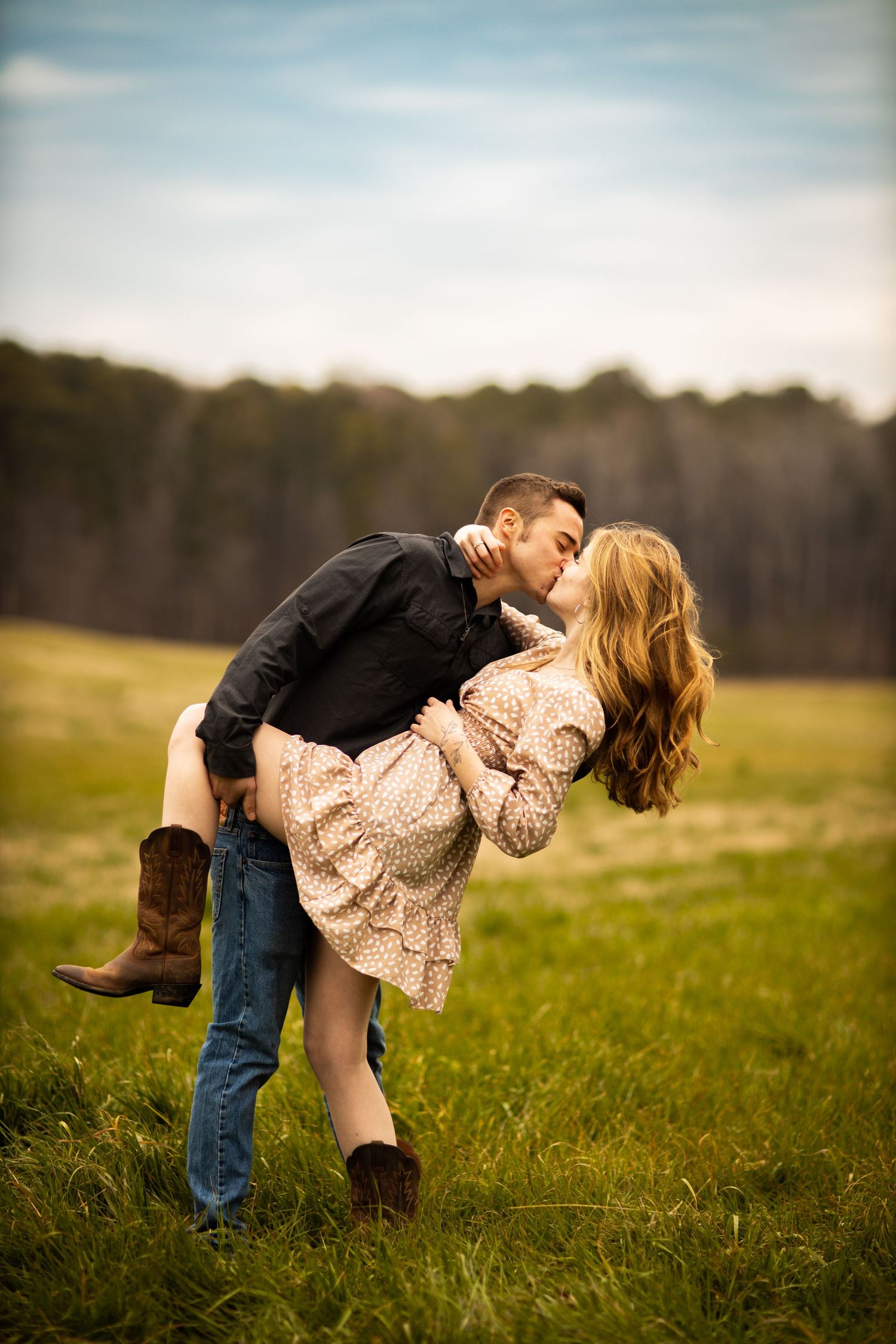 Man lifting woman for a romantic kiss in a lush green field during an outdoor engagement photo session.
