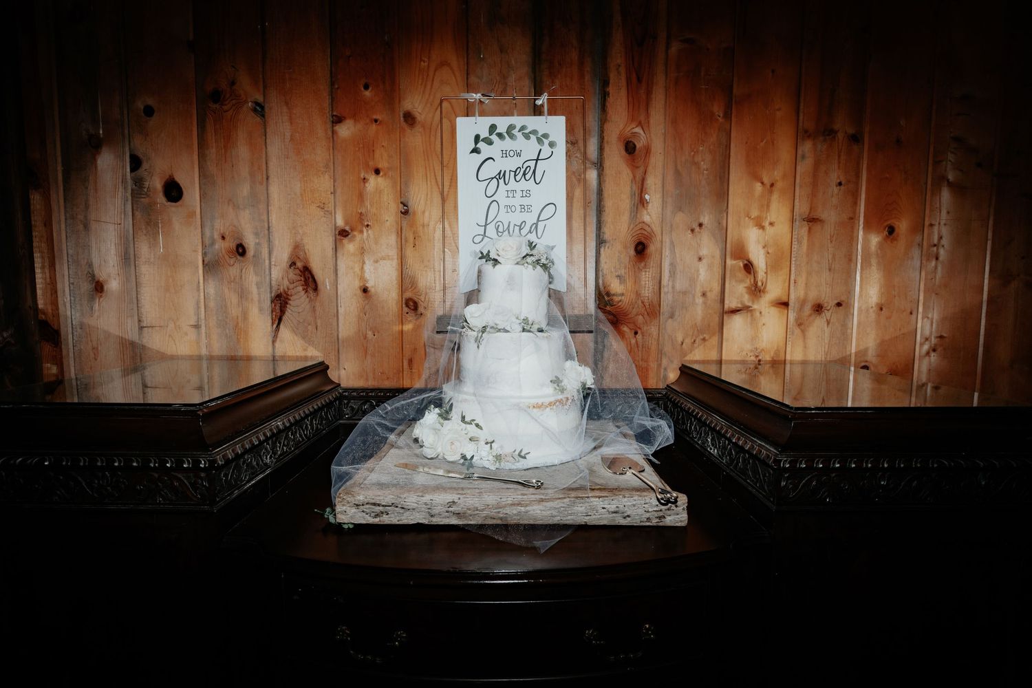 Three-tier white wedding cake displayed on a wooden table with a decorative sign.