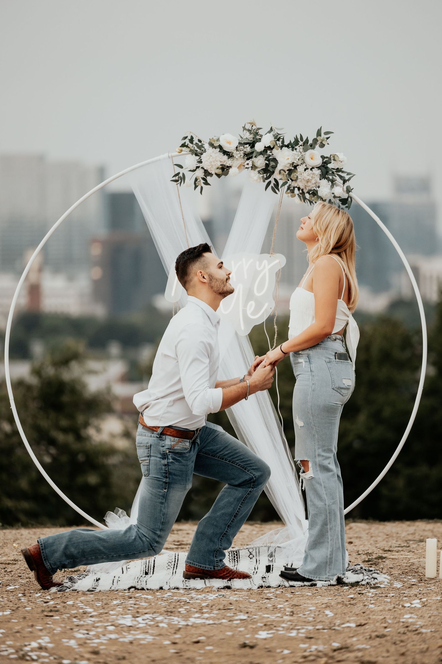 Man proposing on one knee to woman under floral circular arch with city view.