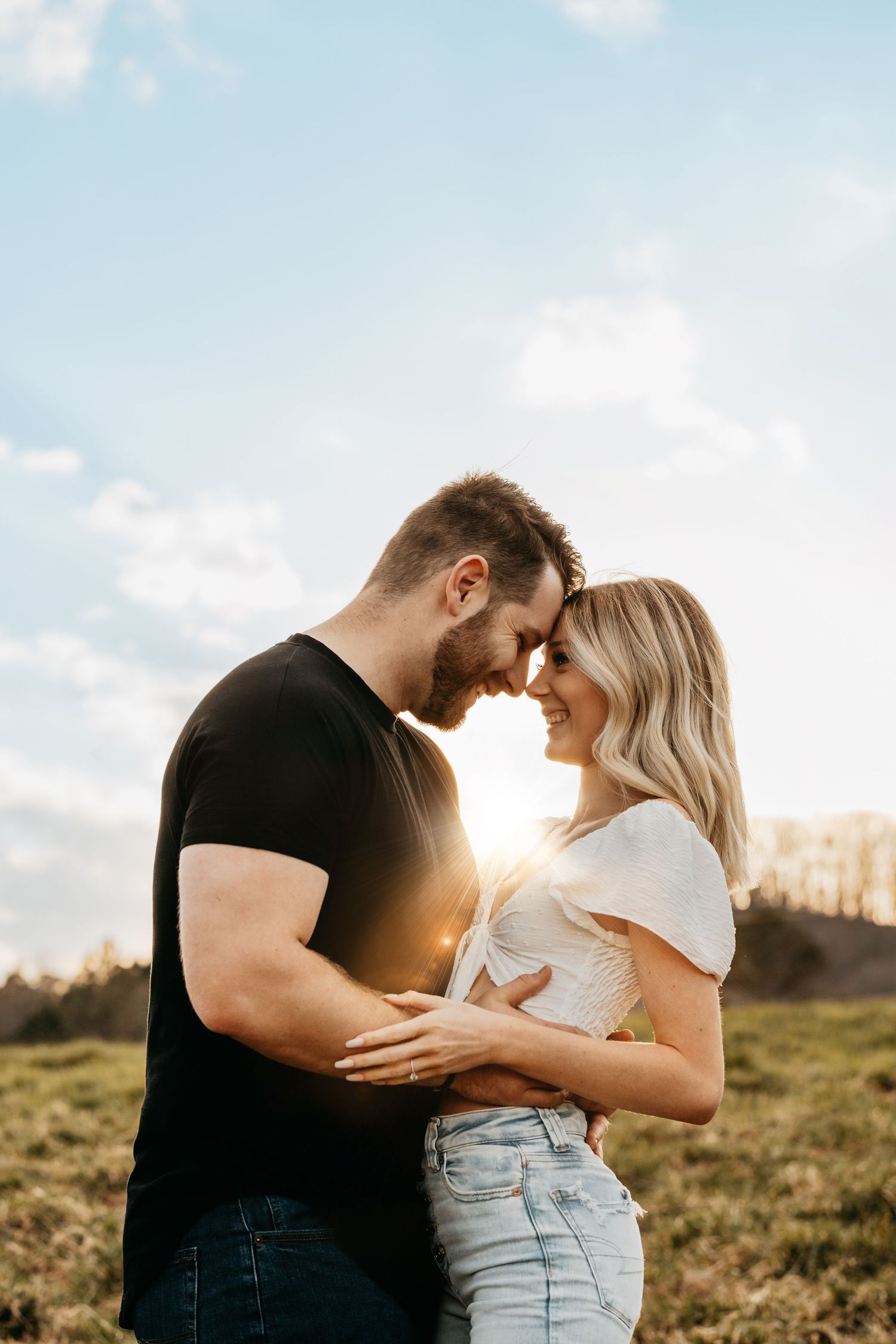 Couple touching foreheads romantically in a sunny field, man in black tee and woman in white crop top and jeans.
