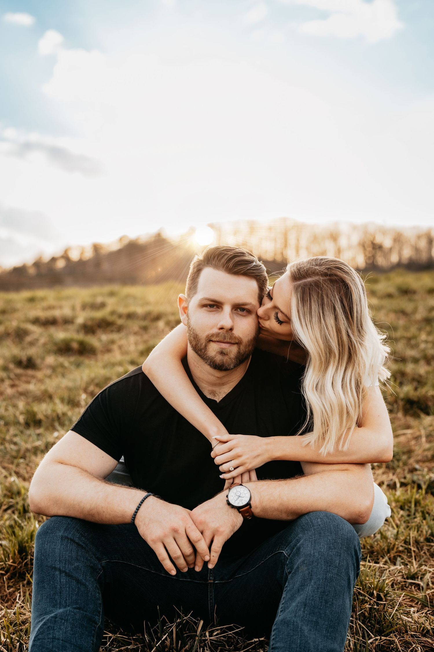 Couple sitting in golden field at sunset, woman embracing man from behind.