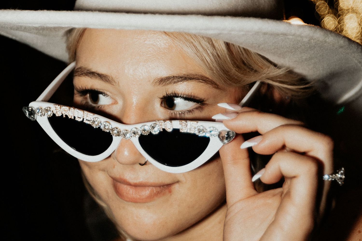 Blonde woman in white cowboy hat peering over rhinestone cat-eye sunglasses smiling.
