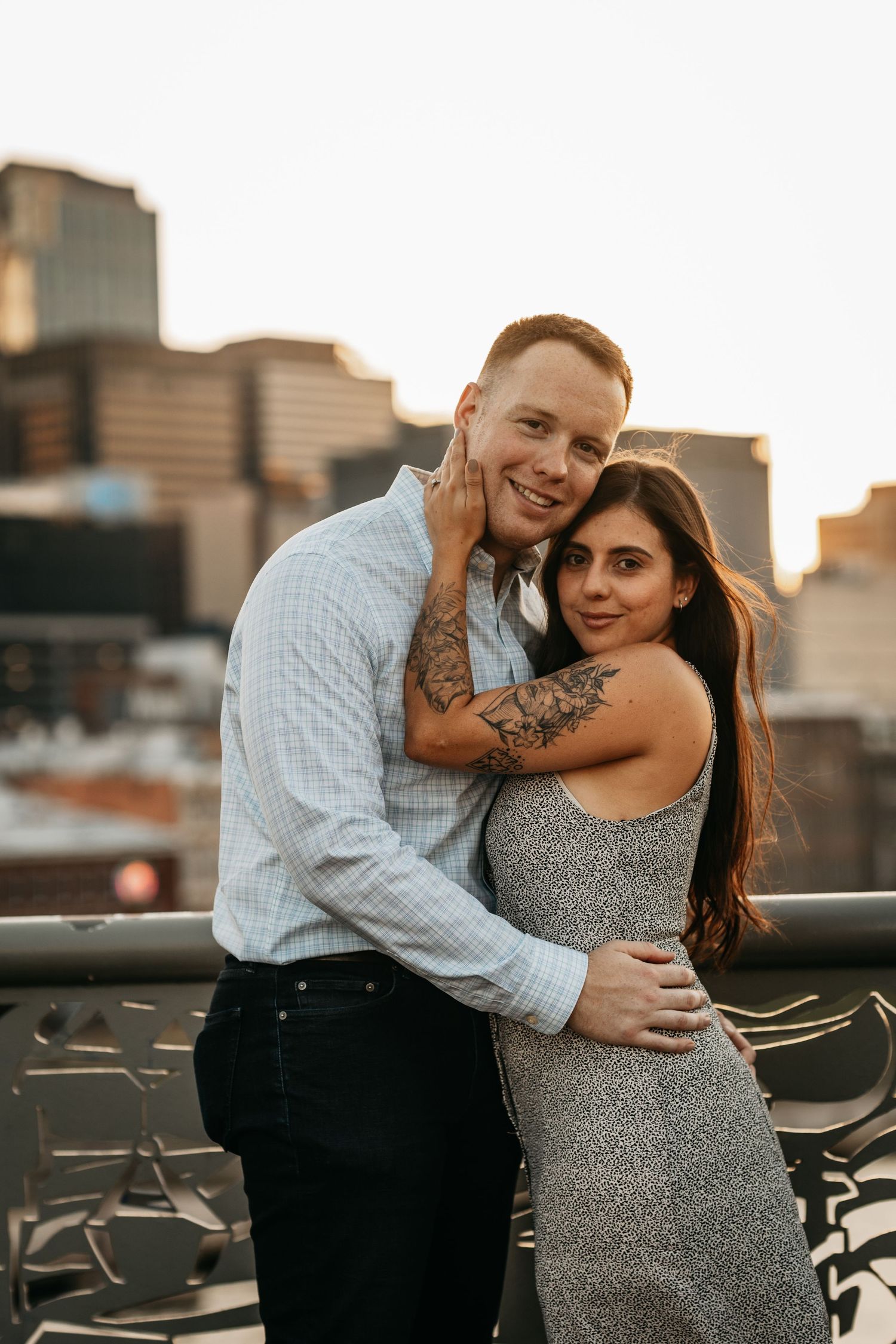 Happy engaged couple embracing on a rooftop with the Nashville skyline glowing at sunset.