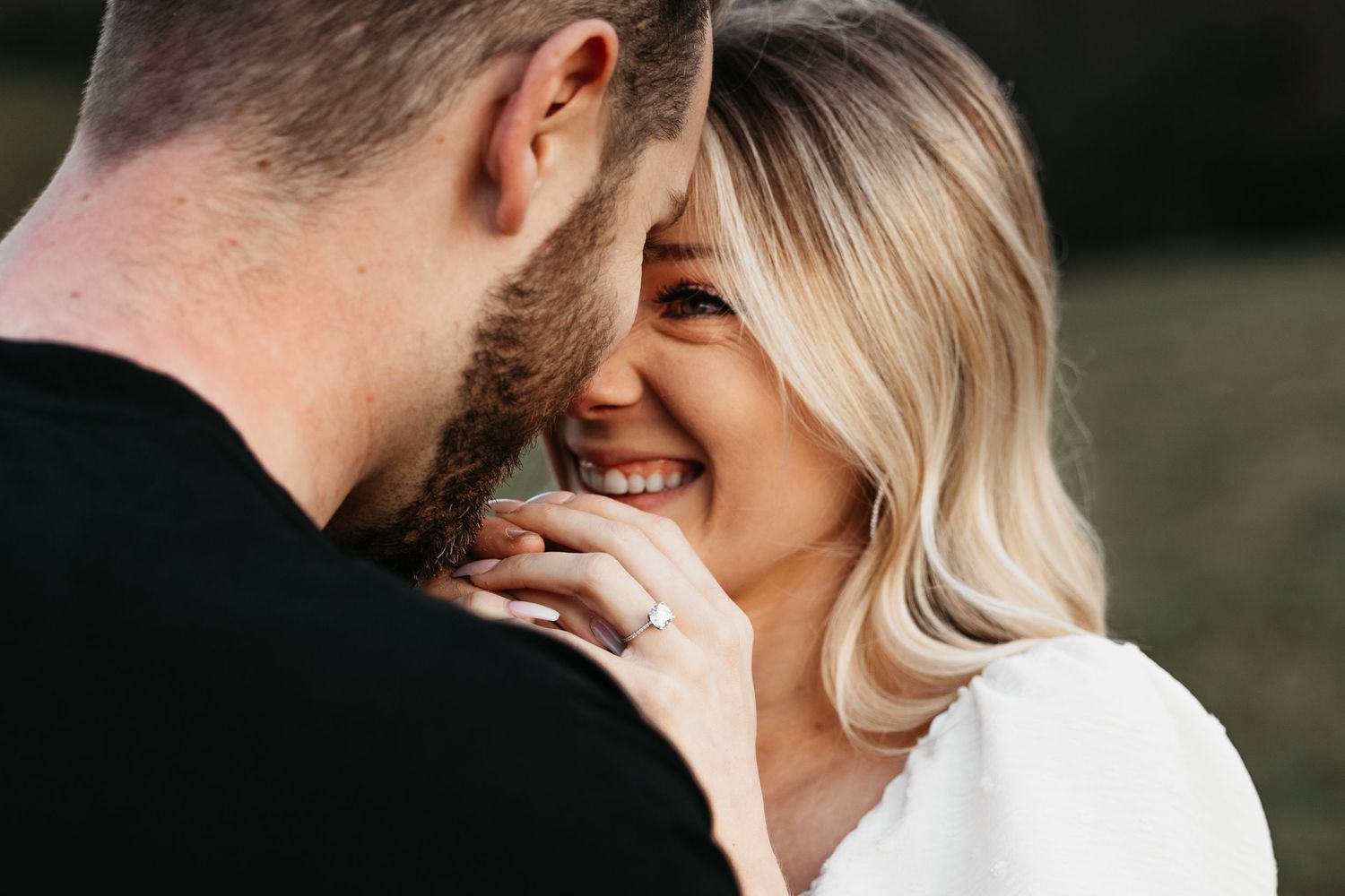 Smiling blonde woman with engagement ring touching bearded mans face closely.
