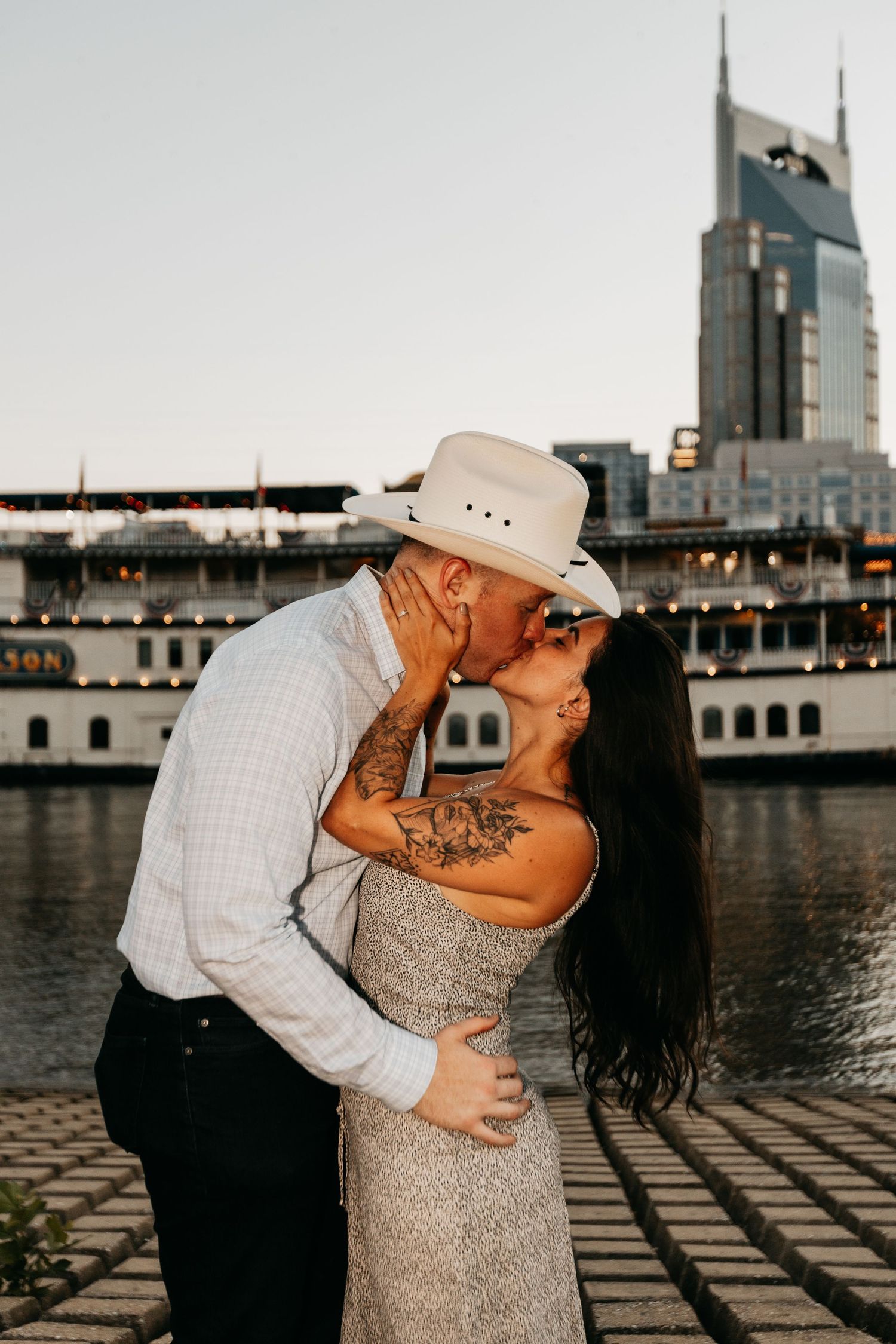 Couple sharing a romantic kiss by the Nashville riverfront with the AT&T building skyline in the background.