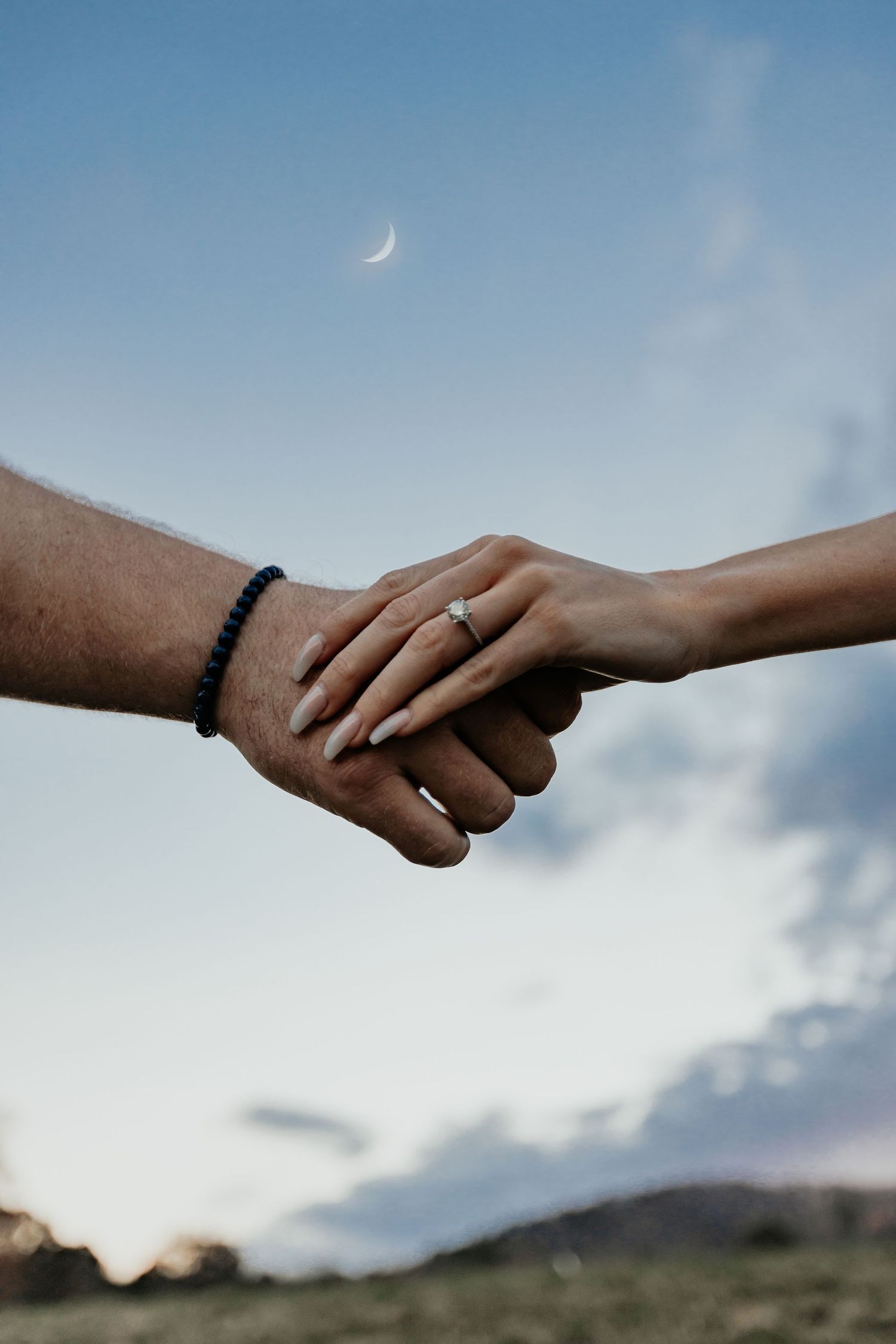 Engaged couple holding hands at dusk with crescent moon visible in sky.