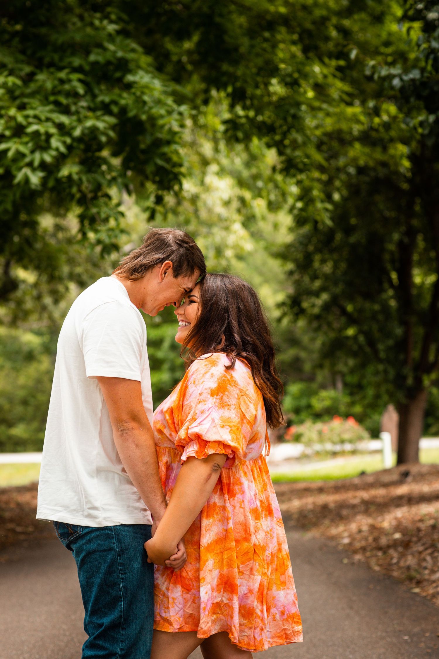 Couple holding hands and smiling at each other on a tree-lined park path during an engagement photo session.