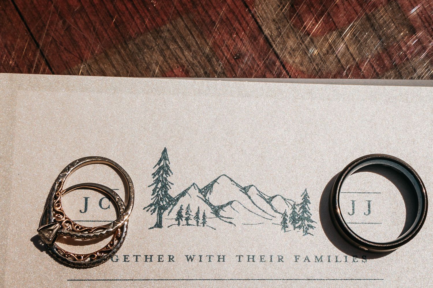 Two wedding rings rest on a mountain-themed wedding invitation with initials J.C. and J.J.