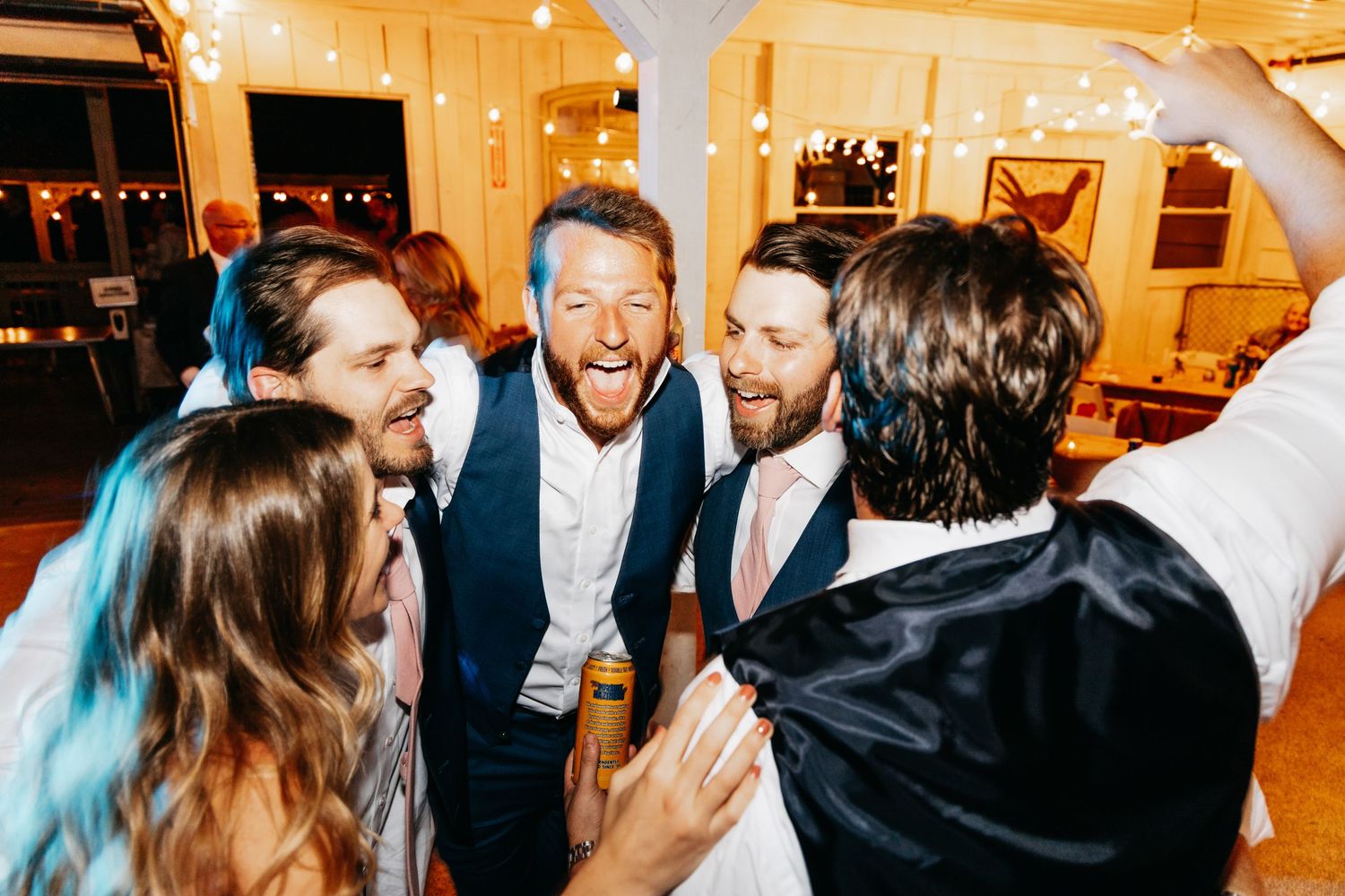 Groom laughing joyfully with groomsmen at a festive wedding reception under string lights.