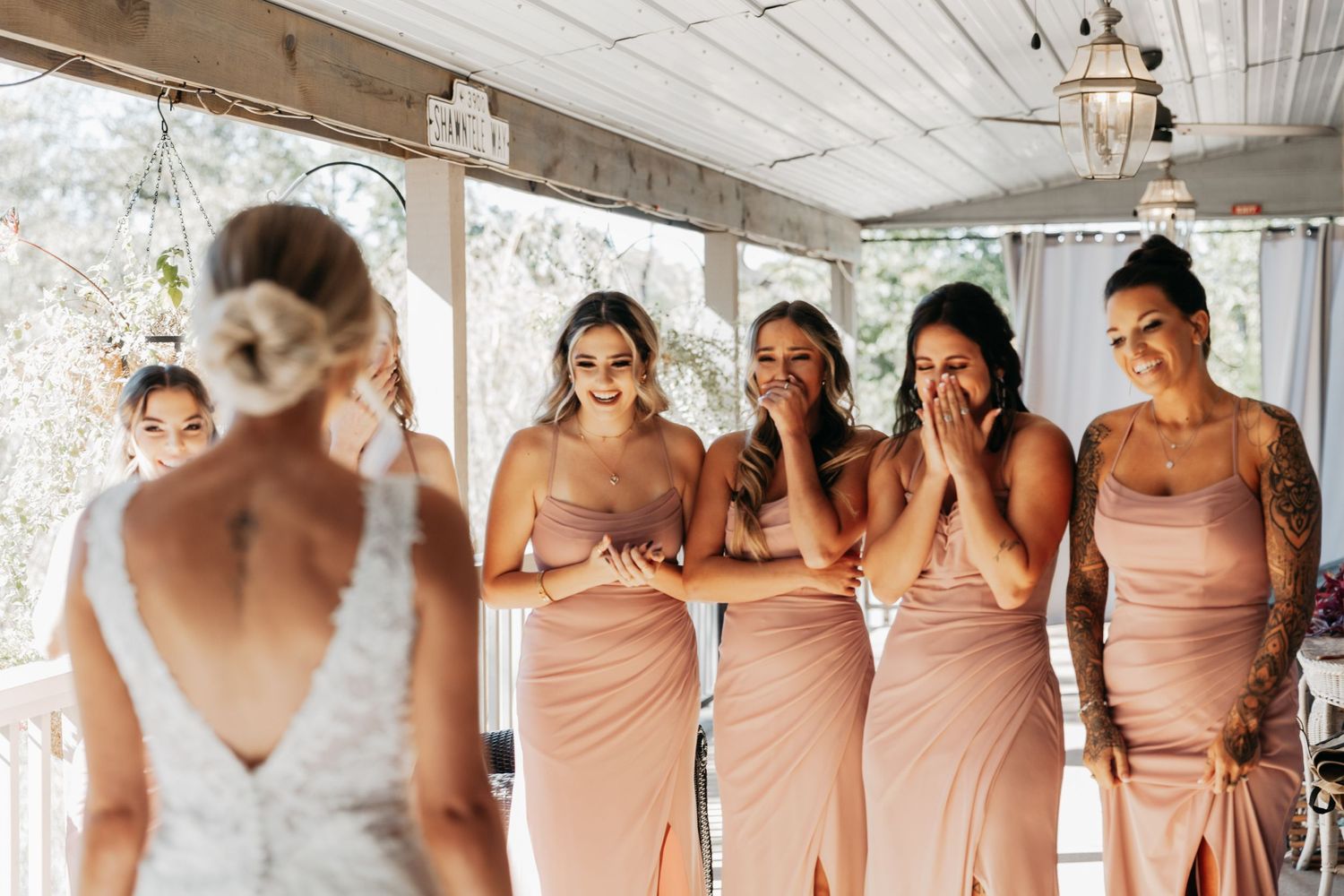 Bridesmaids in blush pink dresses react emotionally during a first look reveal with the bride in a lace wedding gown.