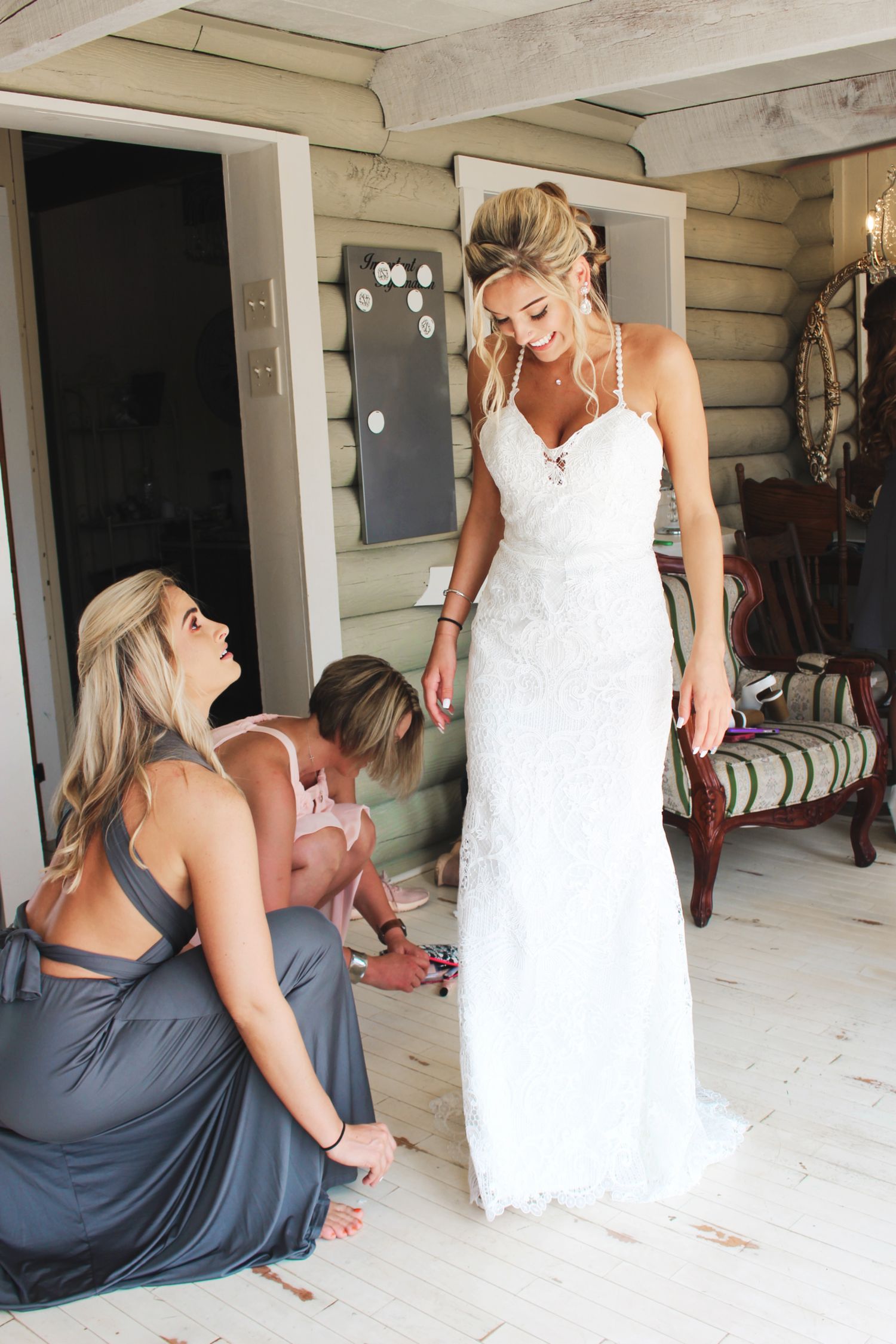 Bridesmaids in gray dresses helping bride into her lace wedding gown indoors.