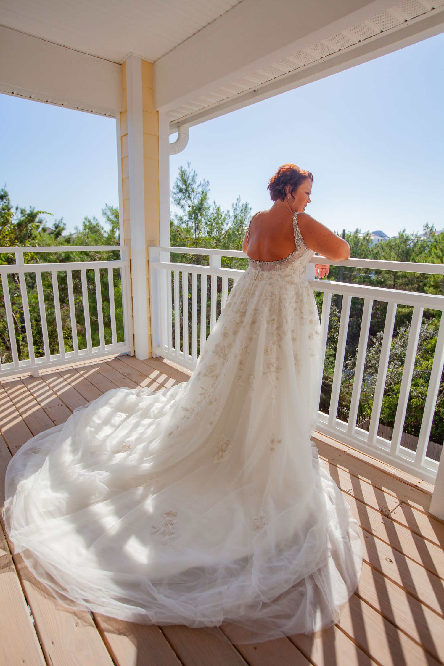 Bride in elegant lace gown with long train standing on a white wooden balcony.