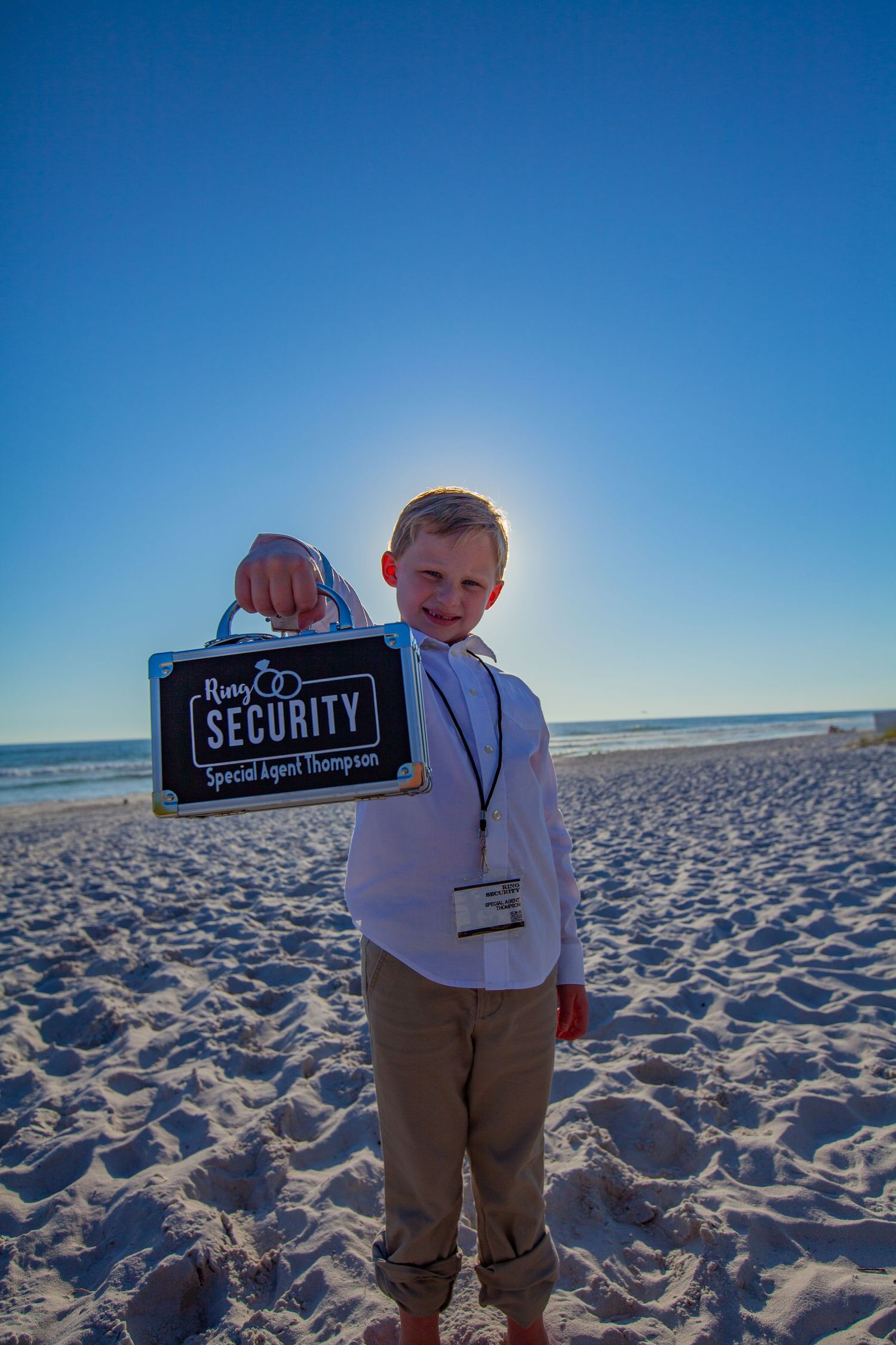 Young boy ring bearer holding a Ring Security briefcase sign on a sunny beach.