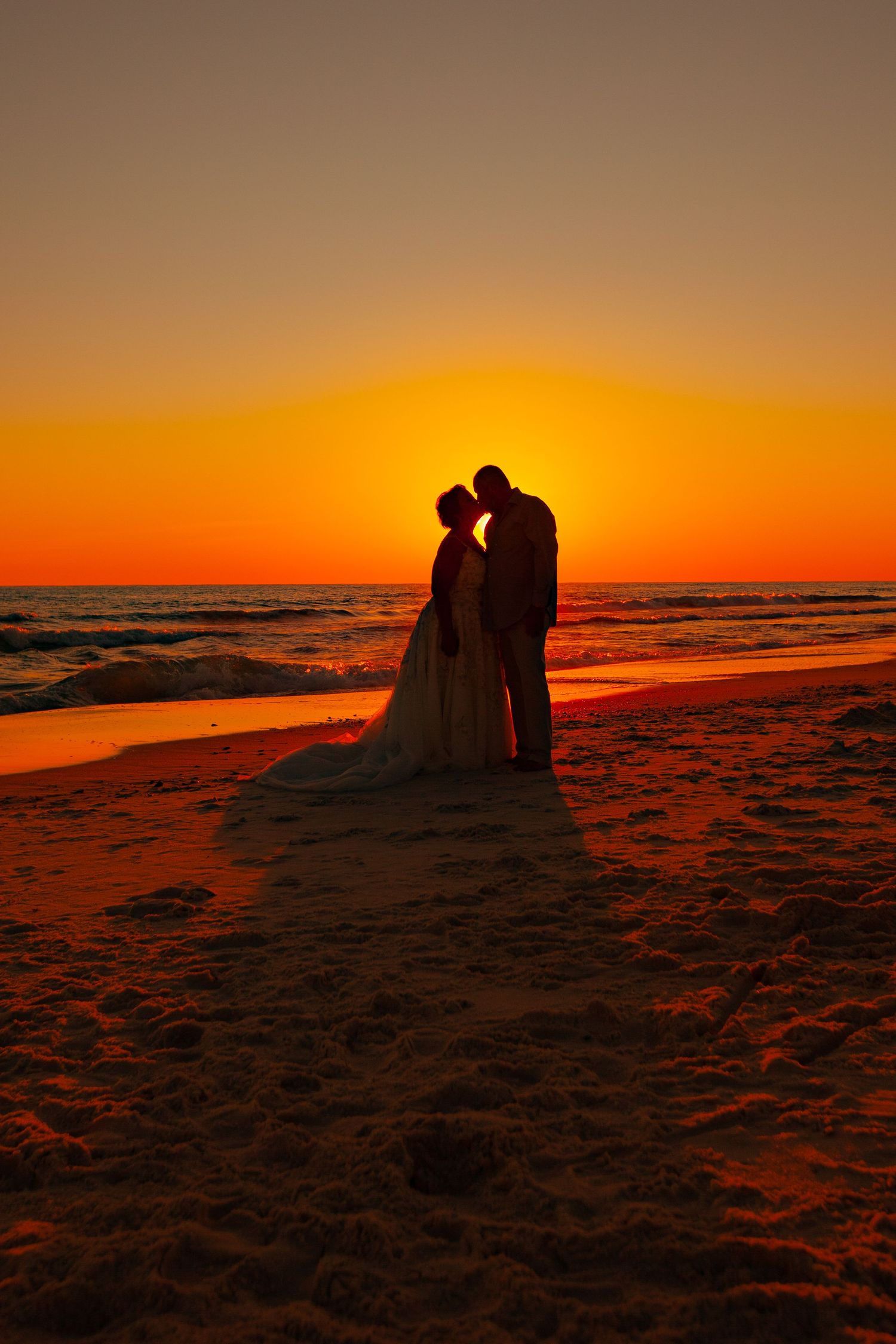 Silhouette of bride and groom kissing on a beach at sunset with vibrant orange sky.