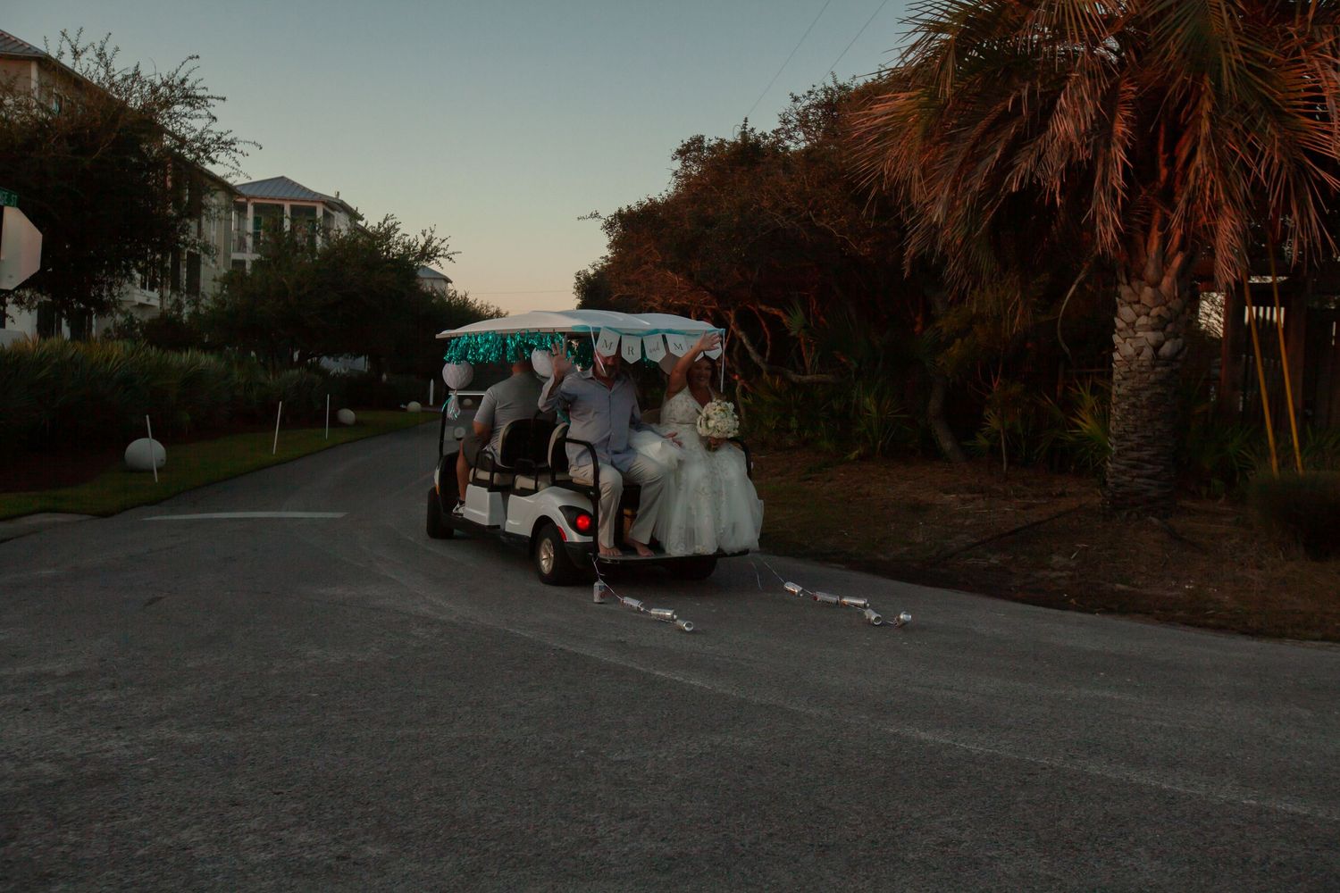 Wedding party riding a golf cart down a tree-lined road at dusk.