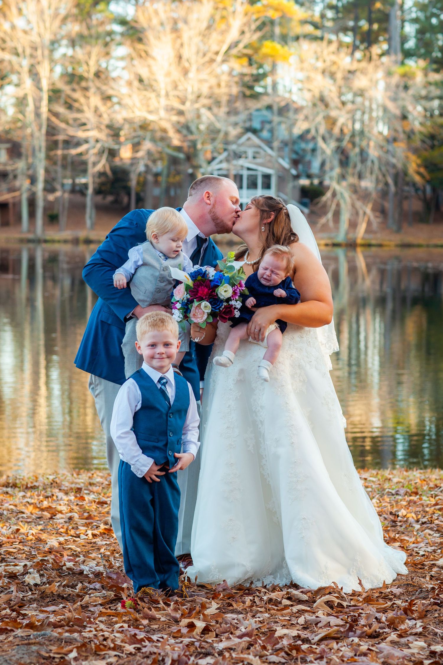 Bride and groom kissing while holding their three young children near a reflective autumn lake.