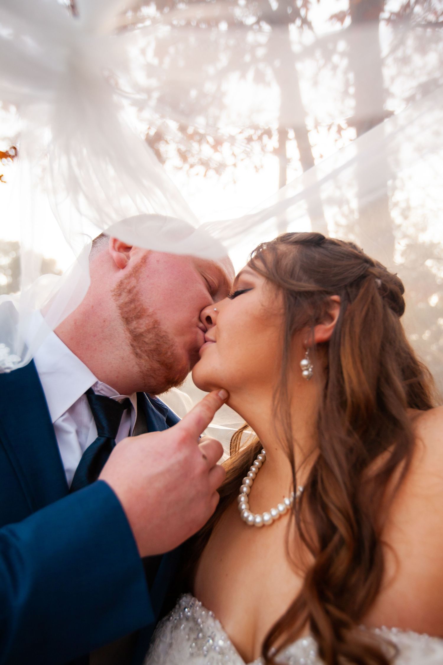 Bride wearing pearl necklace and groom sharing a first kiss under a glowing white floral canopy.