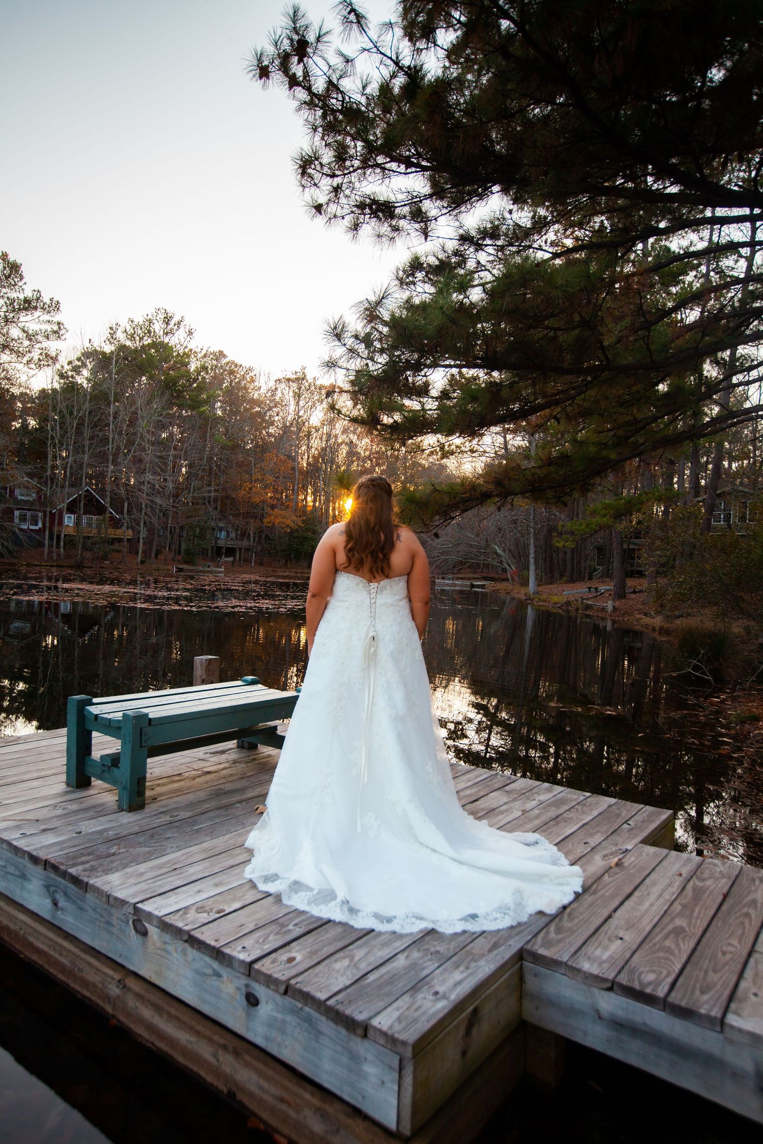 Bride in a white gown standing on a wooden dock at sunset overlooking a peaceful lake.