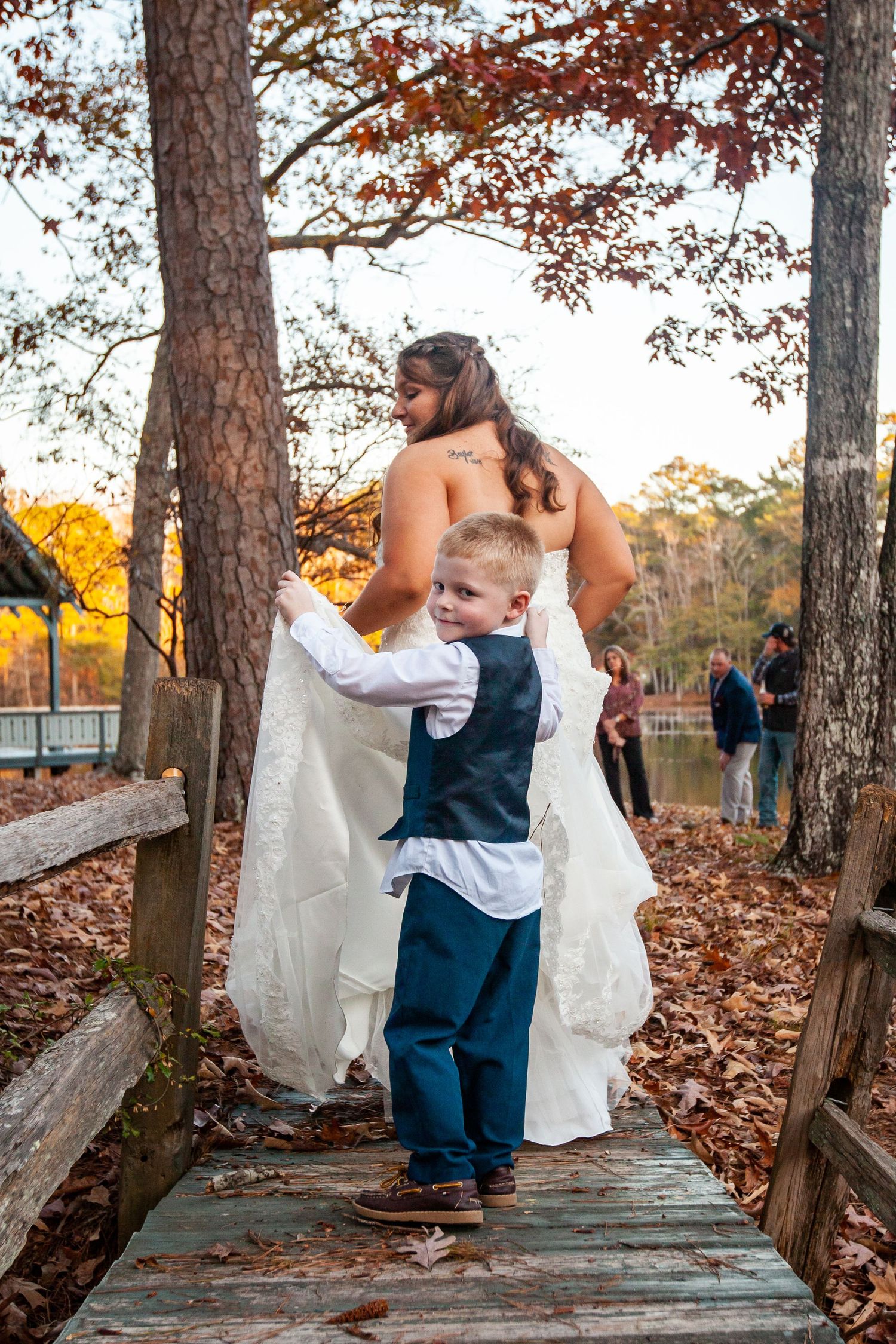 Young boy in a navy vest proudly carrying the bride's train on a rustic wooden bridge.