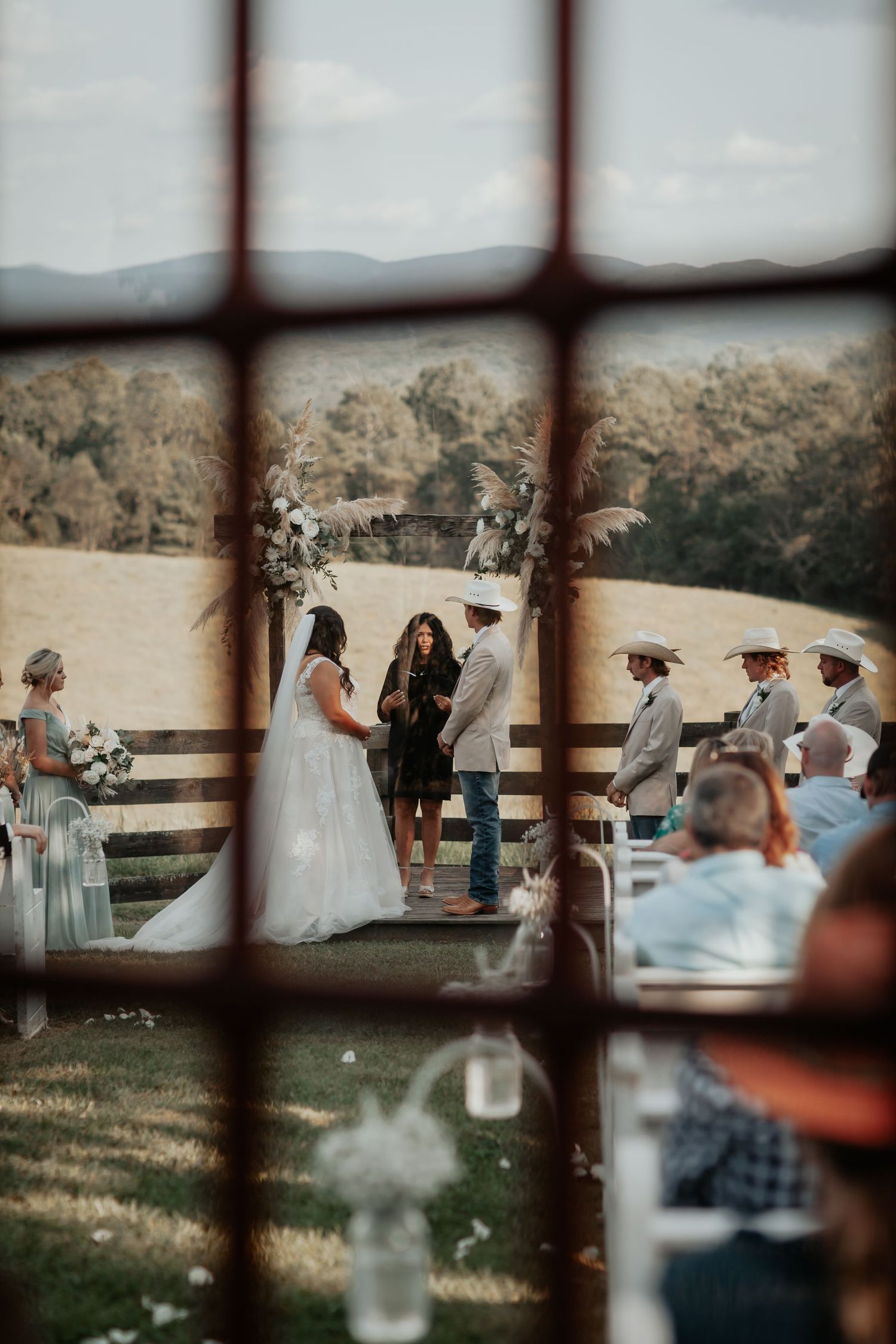 Outdoor rustic barn wedding ceremony viewed through a window with mountain backdrop.