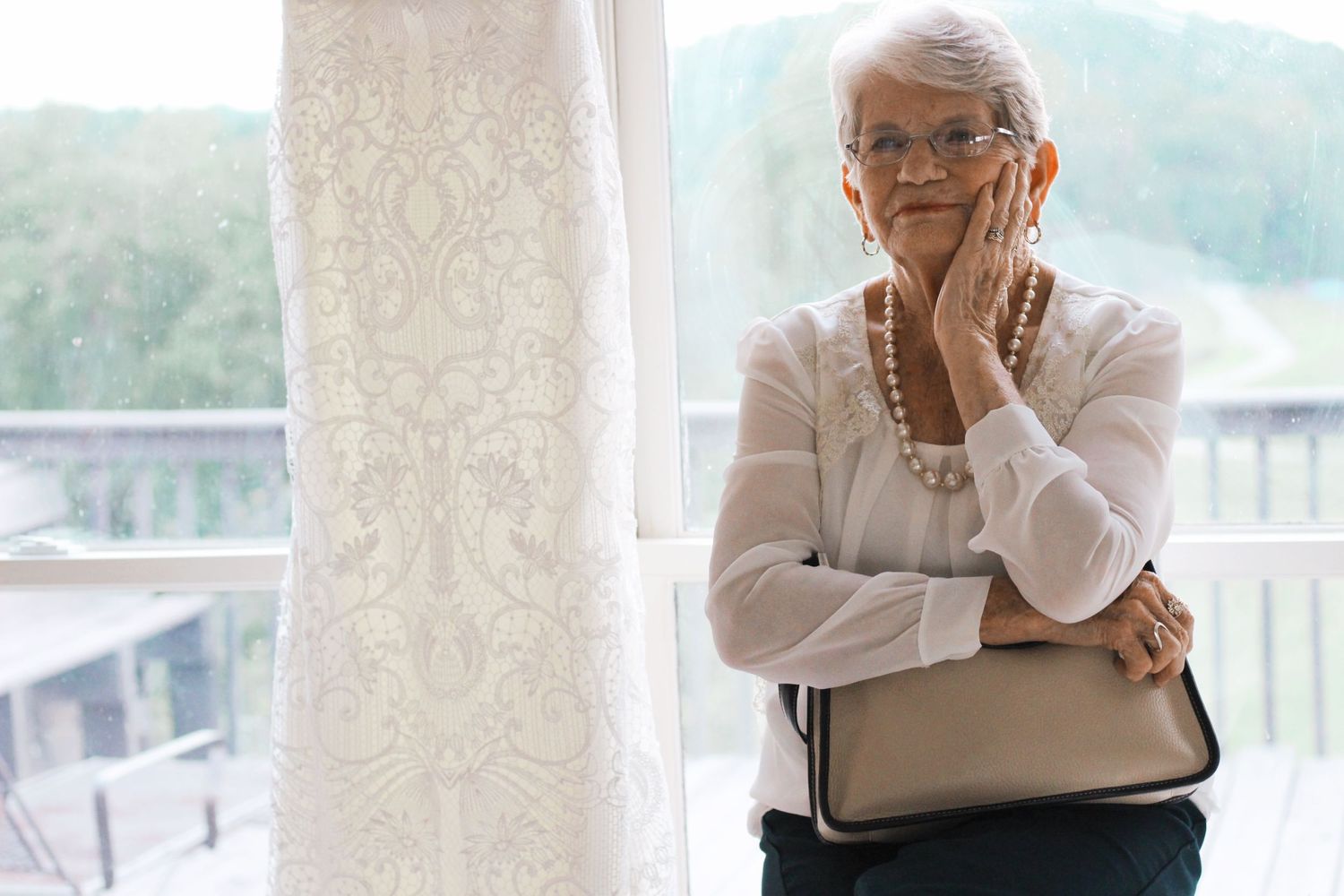 Elegant senior woman in pearls standing beside a wedding dress near a window.