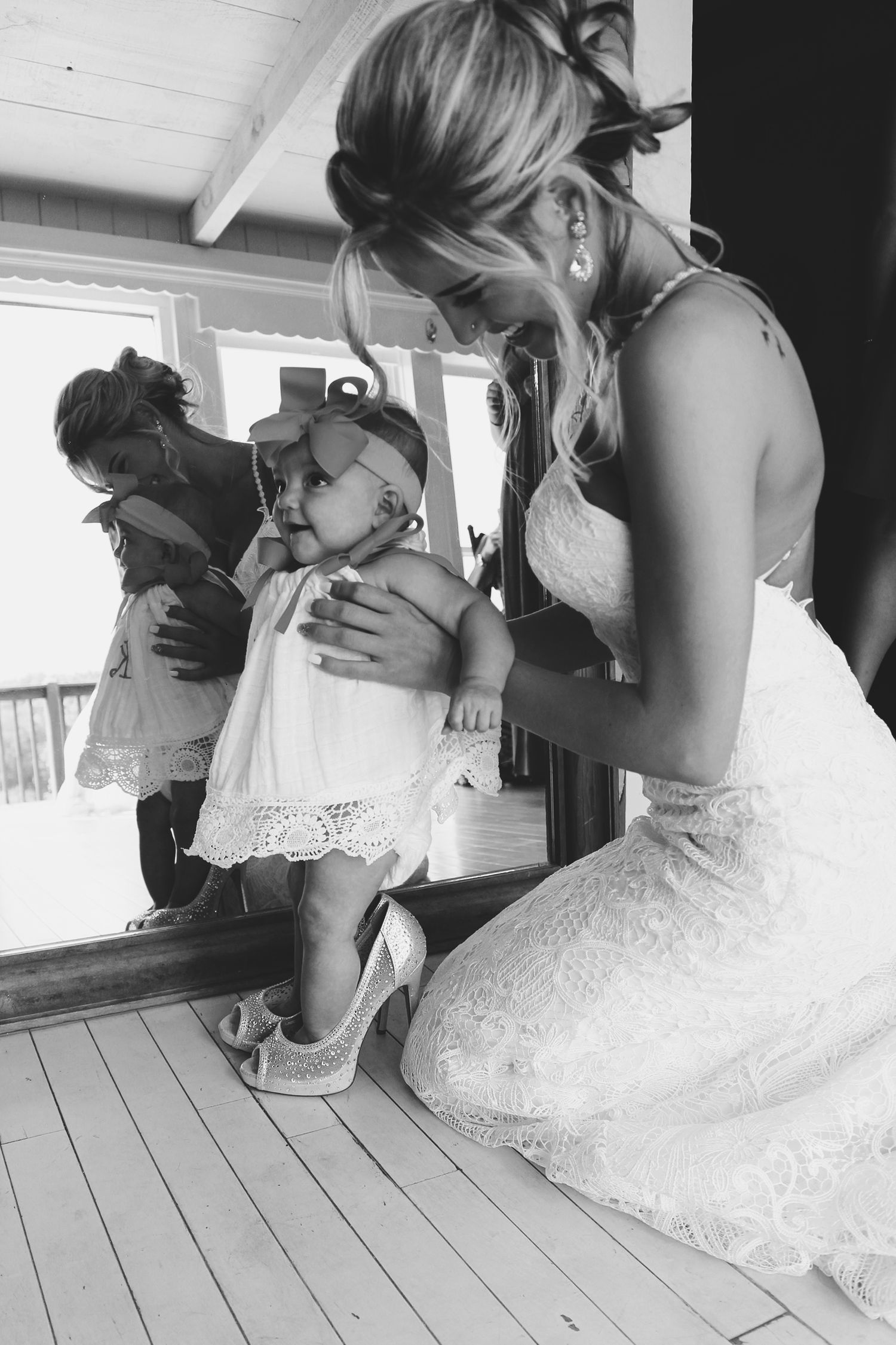 Black and white photo of a bride in a wedding gown kneeling beside a baby girl near a mirror.