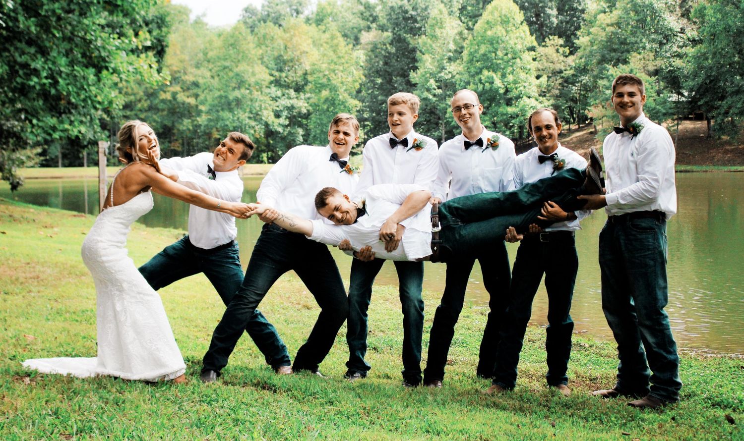 Bride and groomsmen playfully carrying groom horizontally outdoors near a pond.