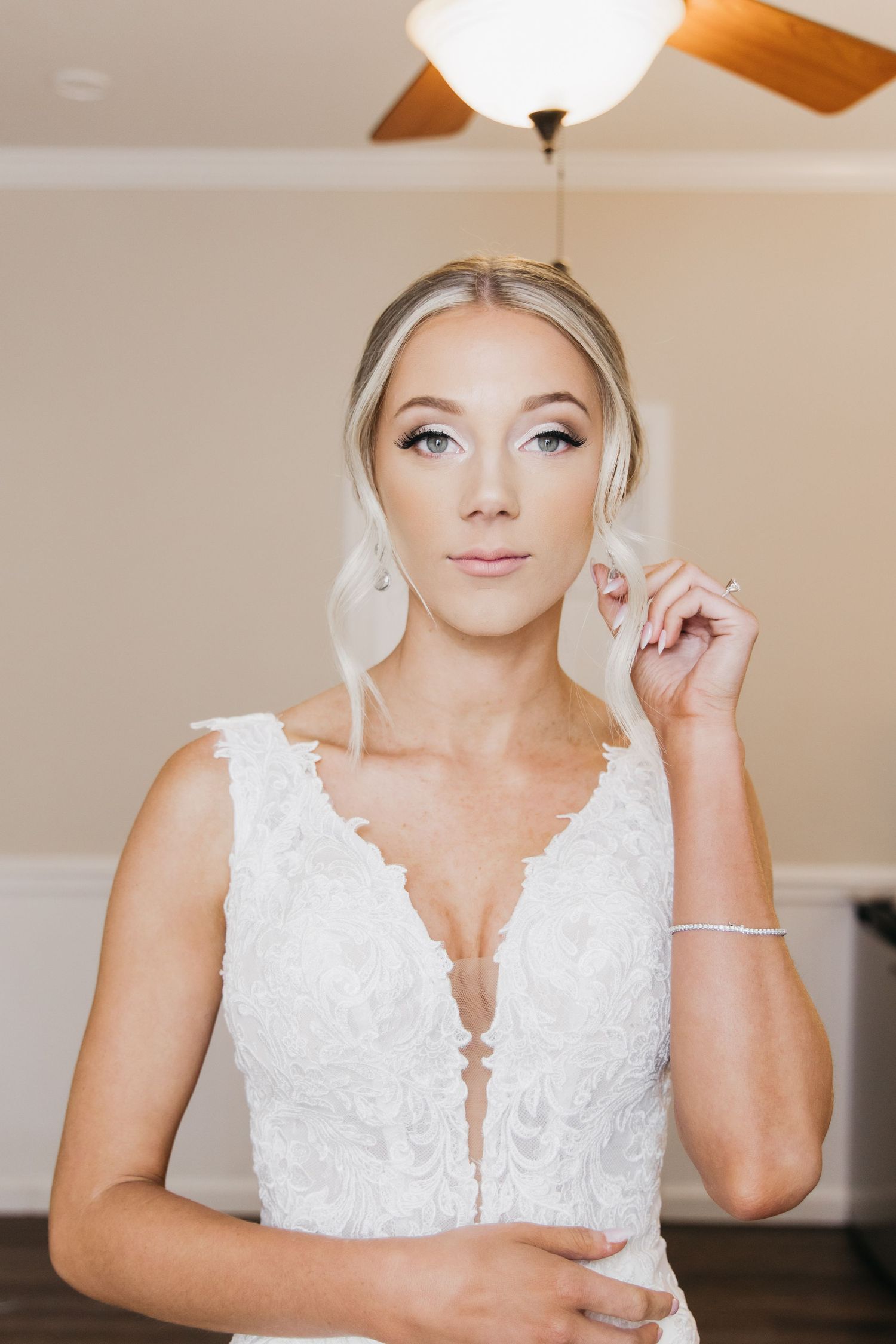 Elegant blonde bride in deep V-neck lace wedding gown adjusting earring indoors.